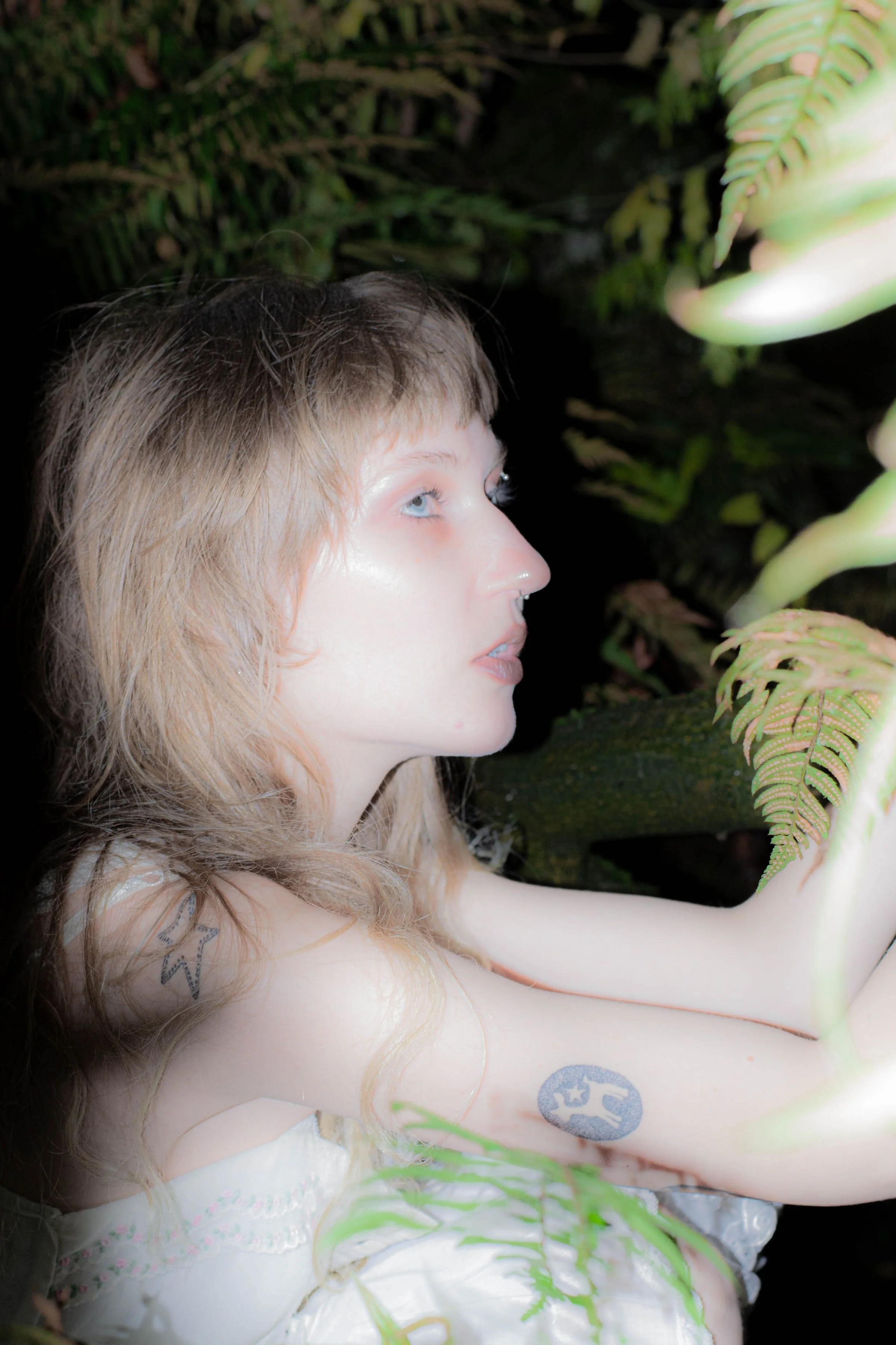 A young woman with light brown hair and light skin looking up with blue eyes, surrounded by green ferns, wearing a white dress with lace details and tattoos on her arm, in a natural setting.