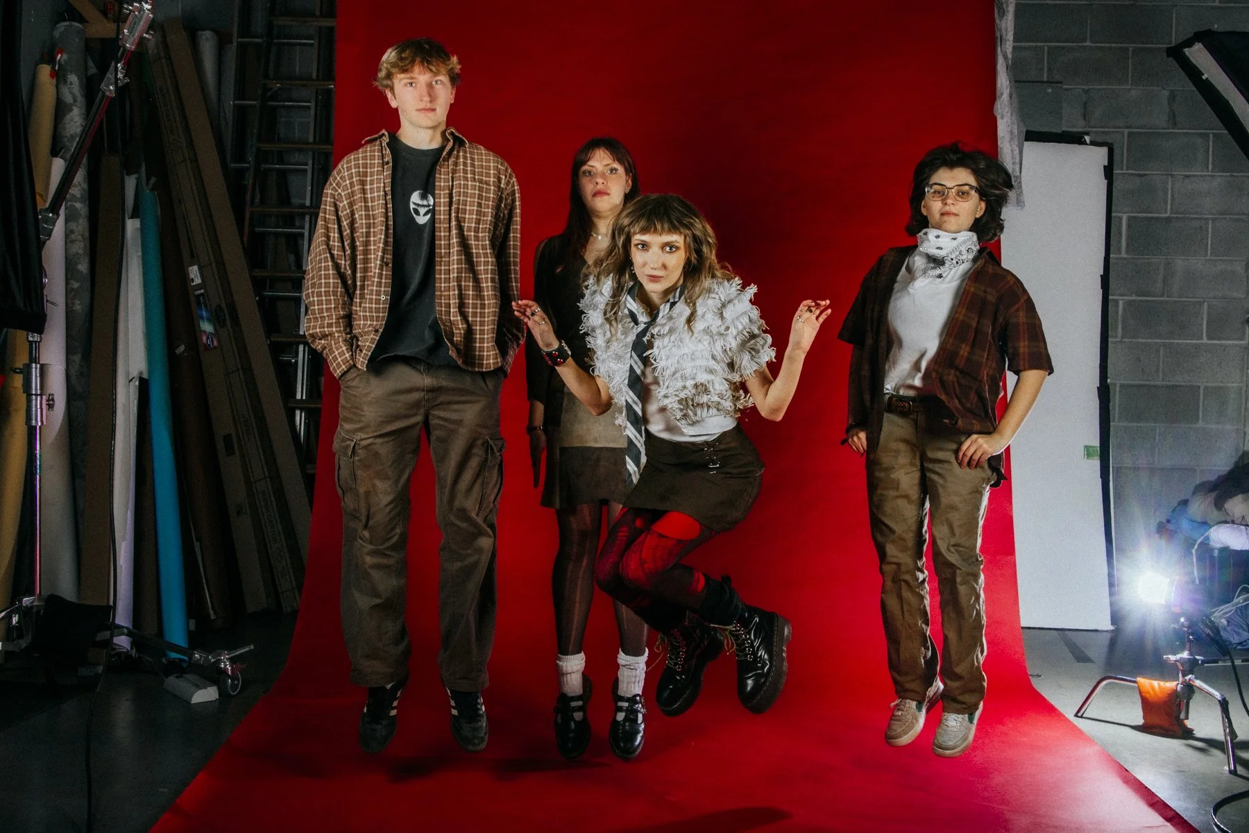 Group of four young people with casual and punk styles posing in front of a red backdrop during a photo shoot, surrounded by photography equipment.