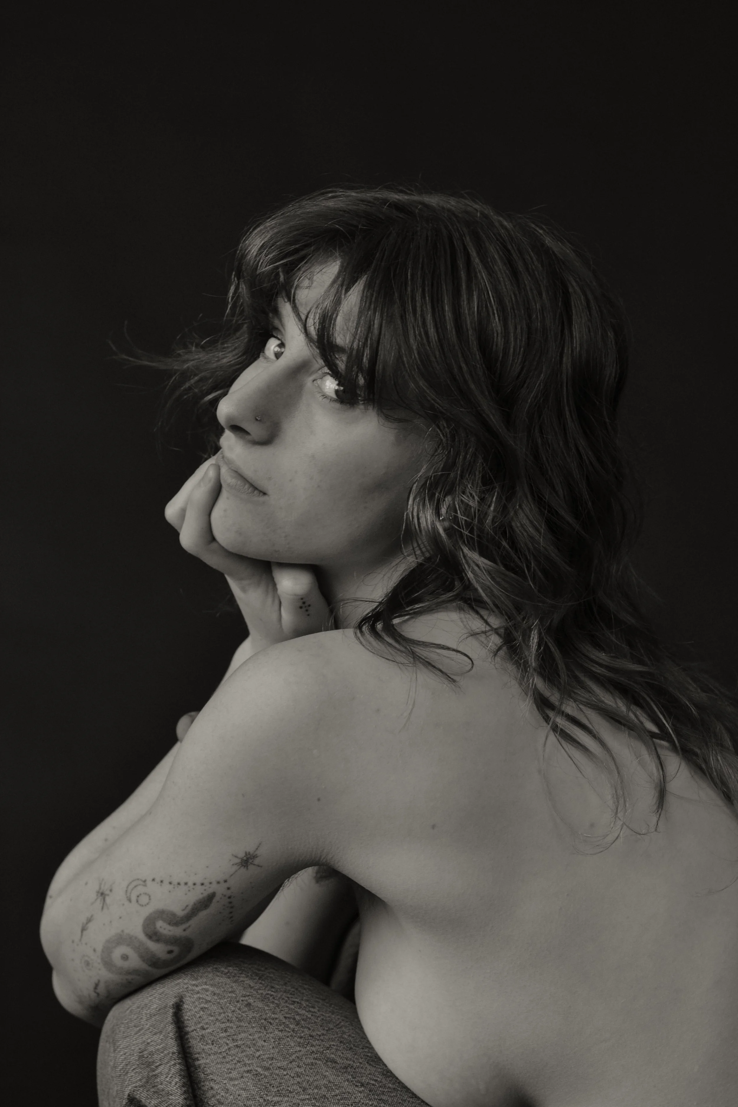 A woman with dark wavy hair and tattoos on her arm, wearing a sleeveless top, resting her chin on her hand against a black background.