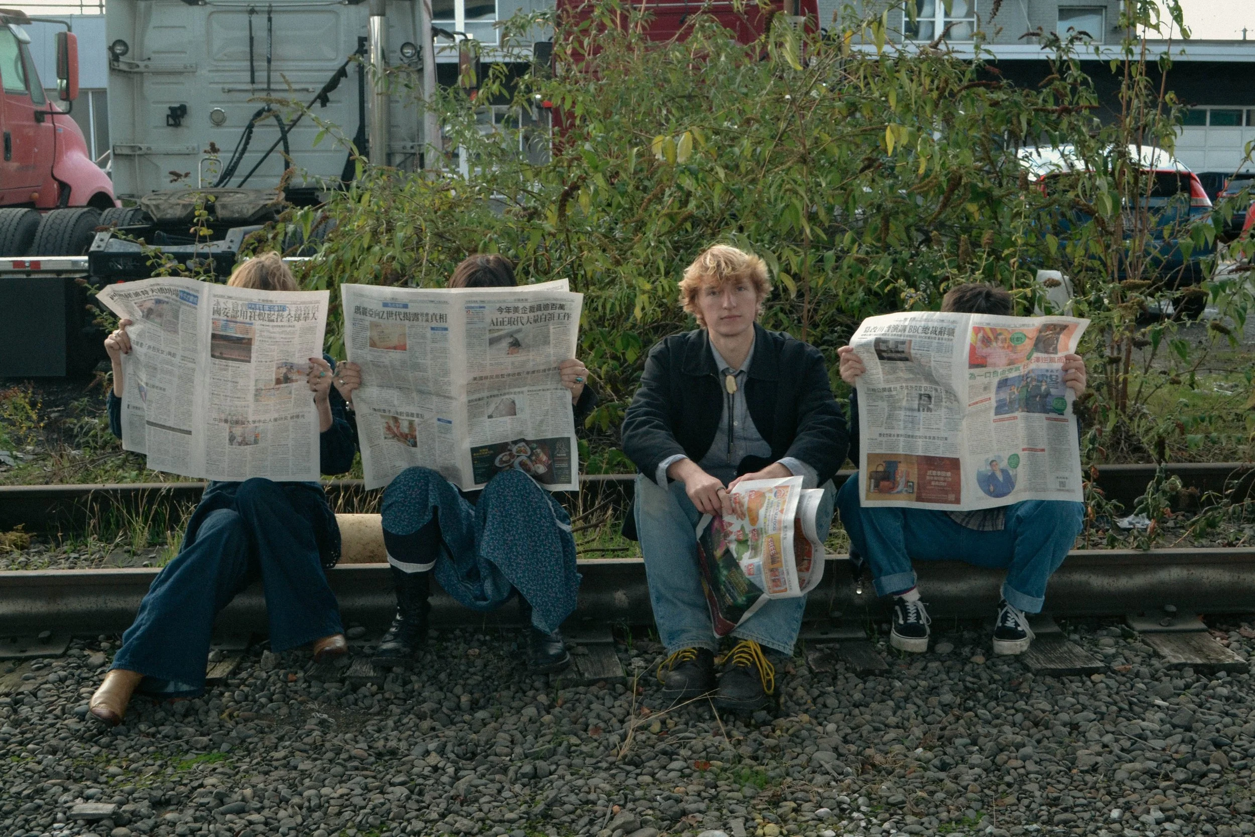 Four people sitting and kneeling on train tracks, holding newspapers, with greenery and parked cars in the background.