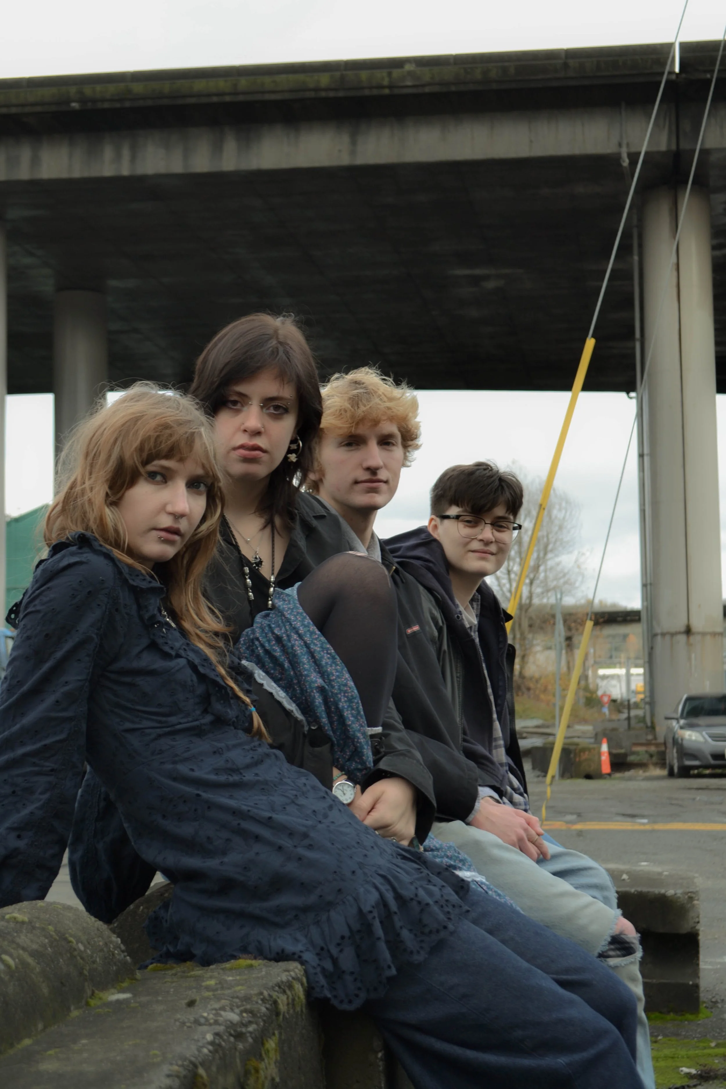 Four young people sitting on a low stone wall outdoors, under a bridge, with urban background and cloudy sky.