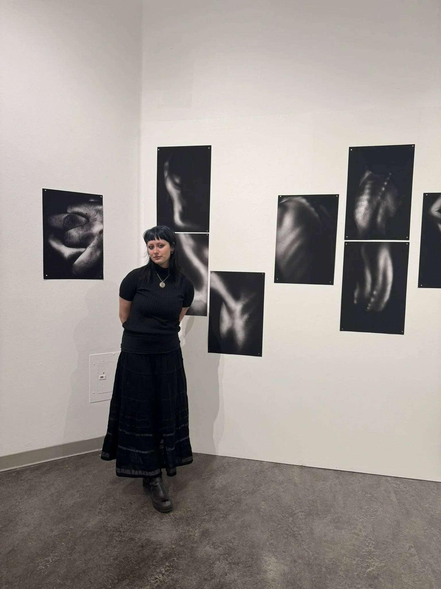 A woman with black hair dressed in a black top and long black skirt standing in front of a white wall adorned with black and white photographs of hands and fists.