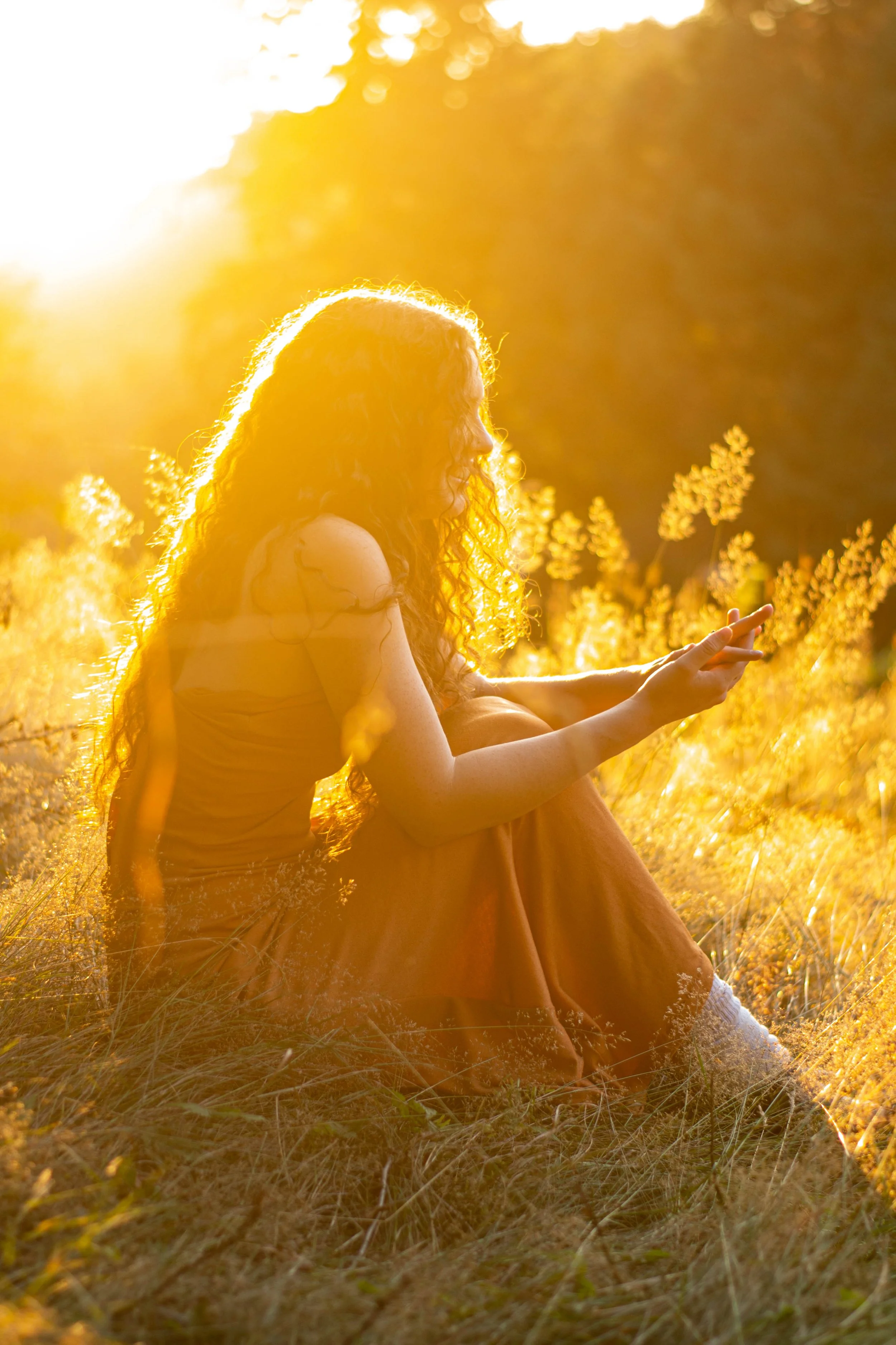 A woman with curly hair sitting on the grass in a field during sunset, bathed in golden light.