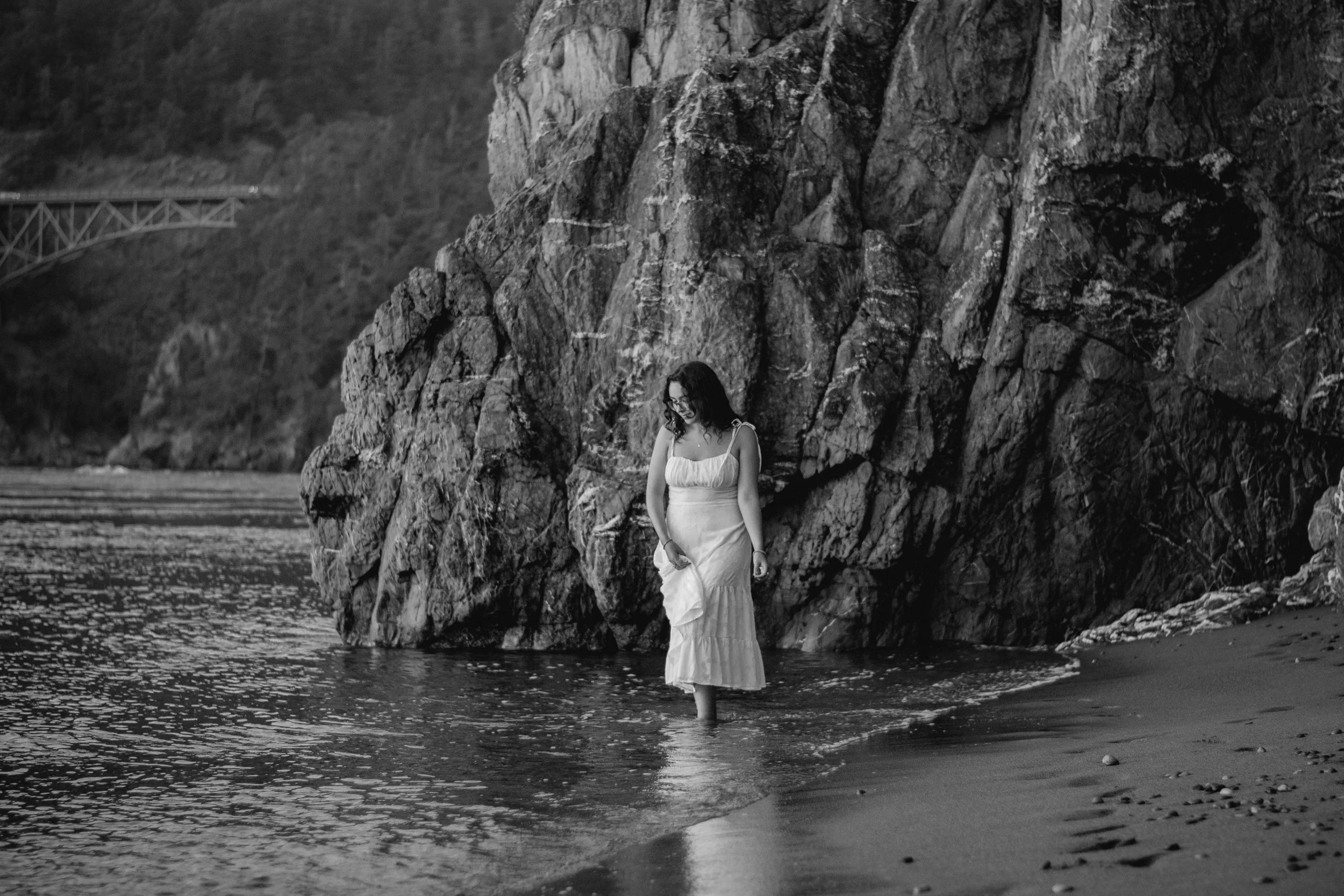 A woman in a long white dress stands in shallow water next to large rocky cliffs, with a bridge visible in the background on the left side of the image.