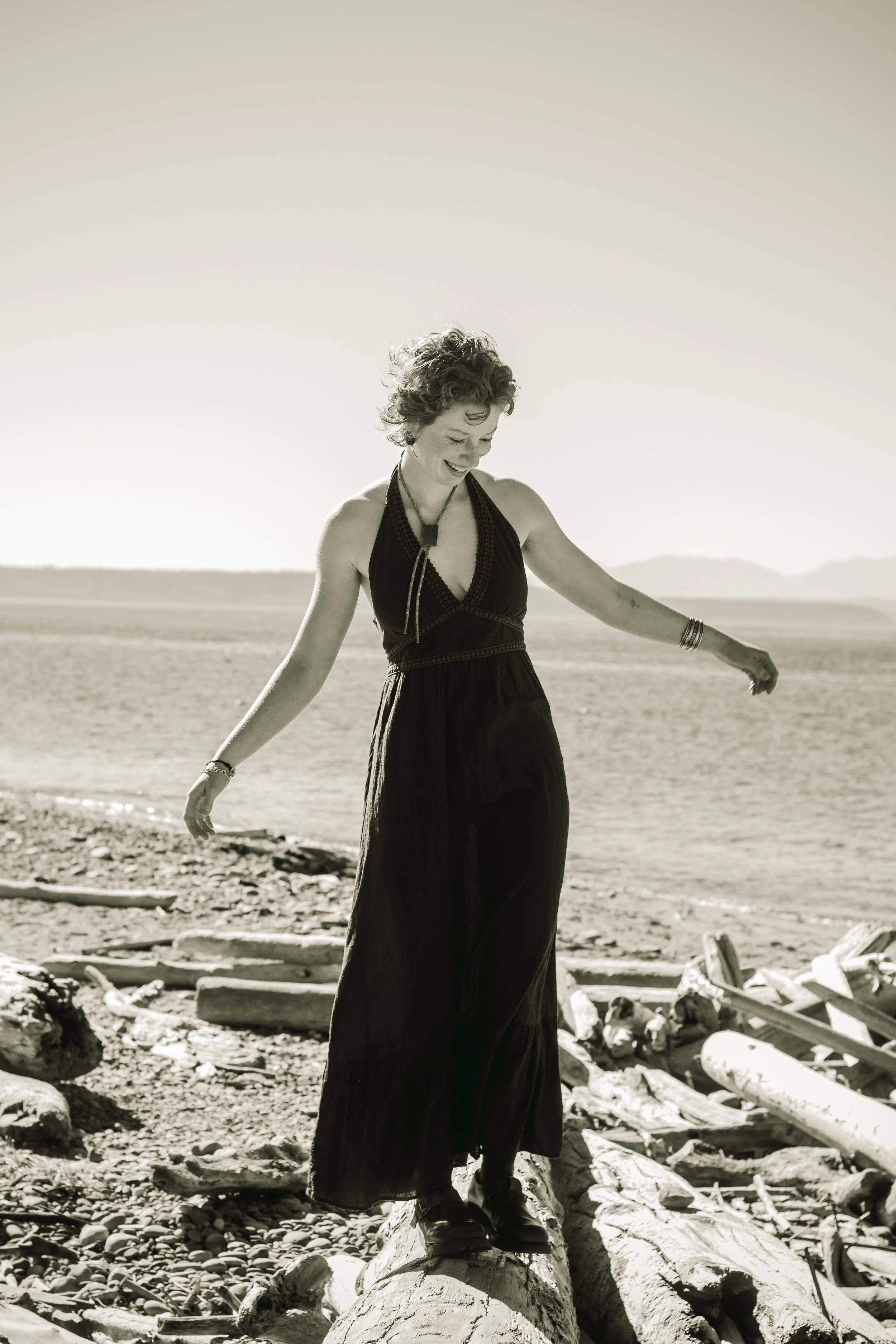 A woman with curly hair, wearing a long black dress, standing on logs at a beach, smiling and looking down, with a distant mountain range in the background.