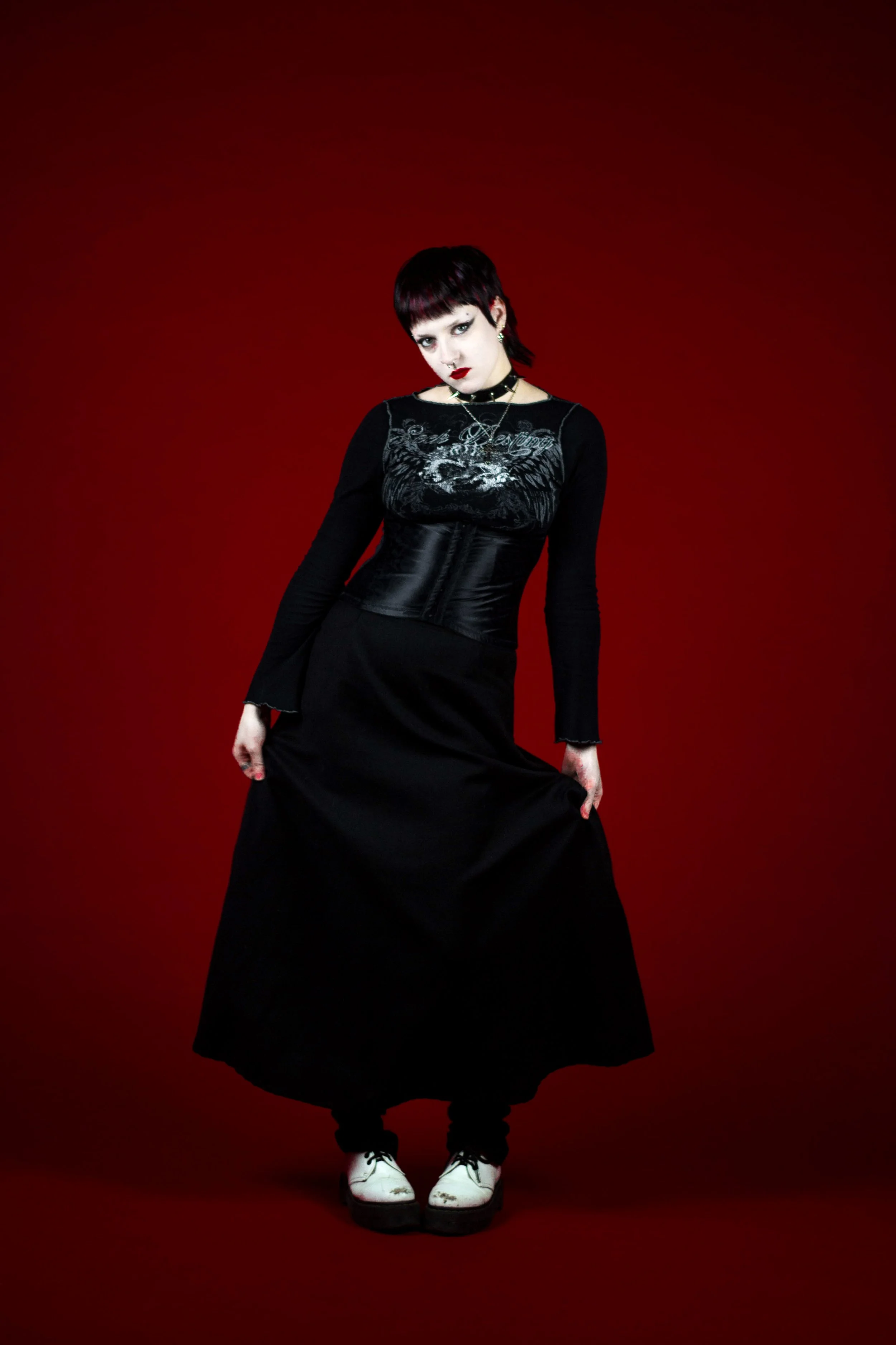 A woman with short dark hair and dark makeup wearing a black graphic long-sleeve top, a long black skirt, white shoes, and multiple jewelry items against a red background.