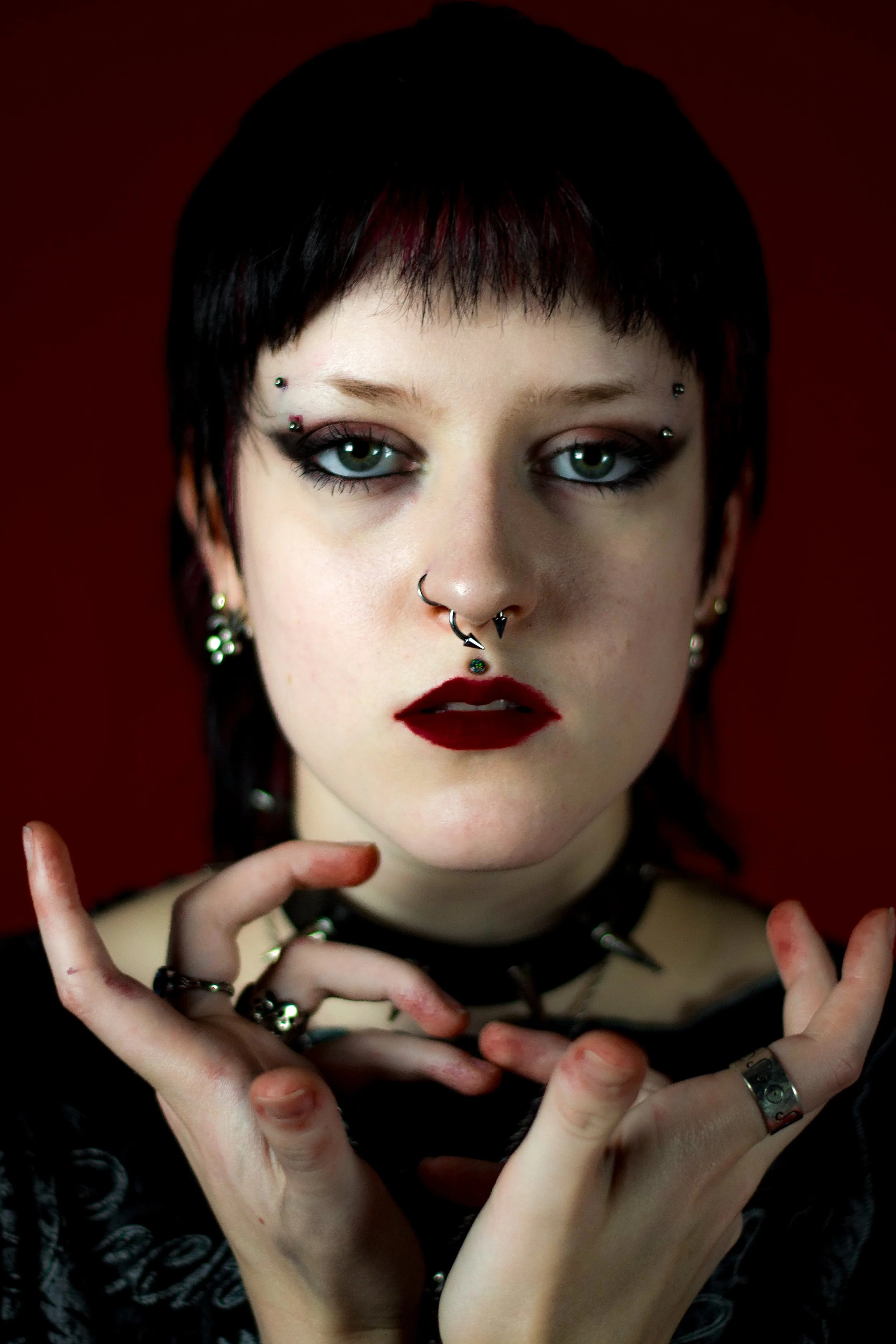 A person with short dark hair, green eyes, and makeup, wearing multiple facial piercings, black jewelry, and a black choker with spikes, holding their fingers in a delicate pose against a dark red background.