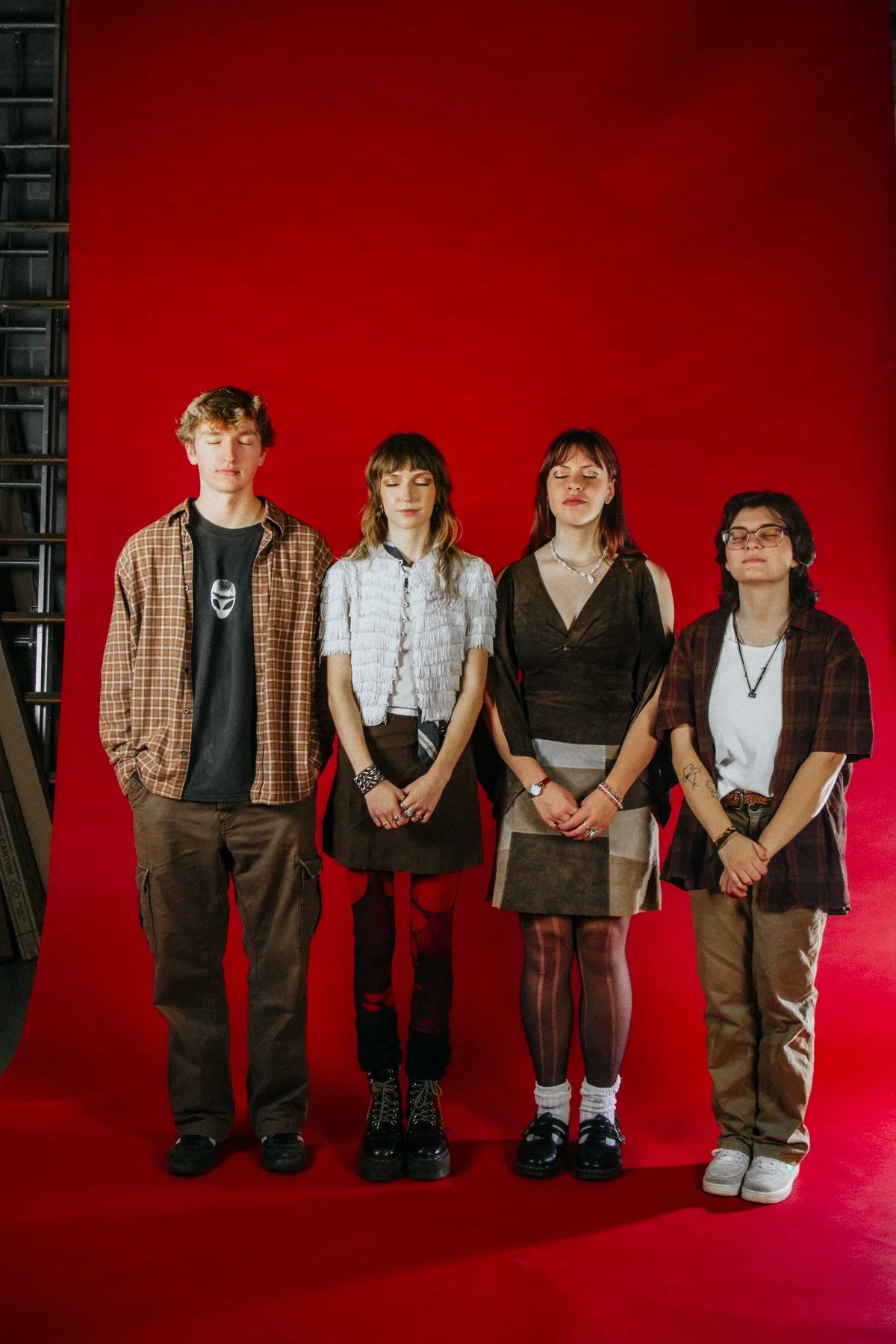 Four young people standing with eyes closed against a red background, with the first person wearing a checkered shirt, the second in a white textured shirt, the third in a black top and skirt, and the fourth in a white shirt with a checkered over shi