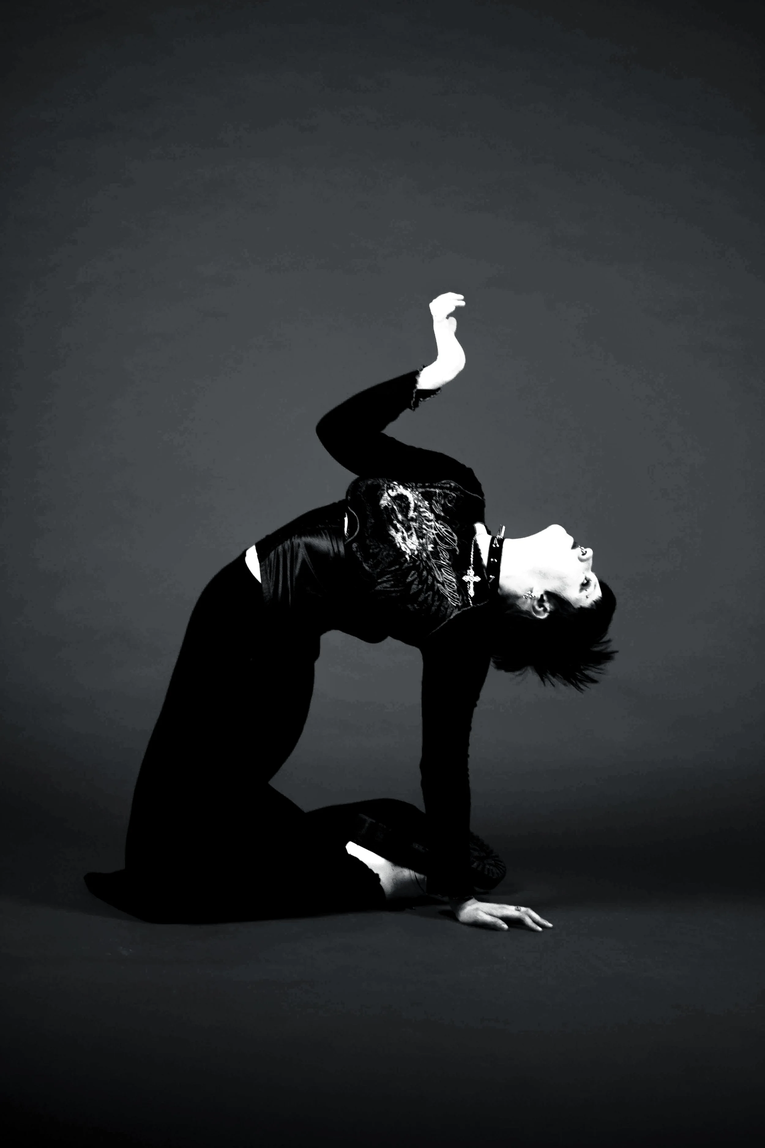 A black and white photo of a woman performing a yoga pose on a dark background. She is on her hands and one knee, with her other arm raised and her head tilted back, smiling.