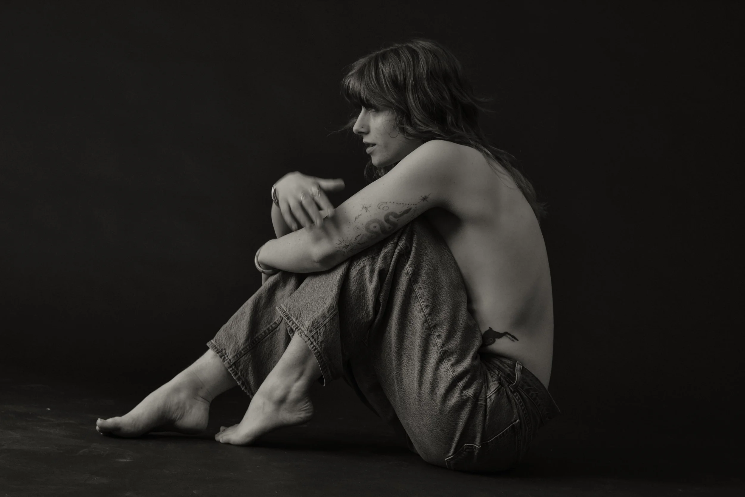 A black and white photo of a woman sitting on the floor with her arms wrapped around her knees, wearing denim jeans and a shirt draped over her shoulders, with tattoos on her left arm and a contemplative expression.