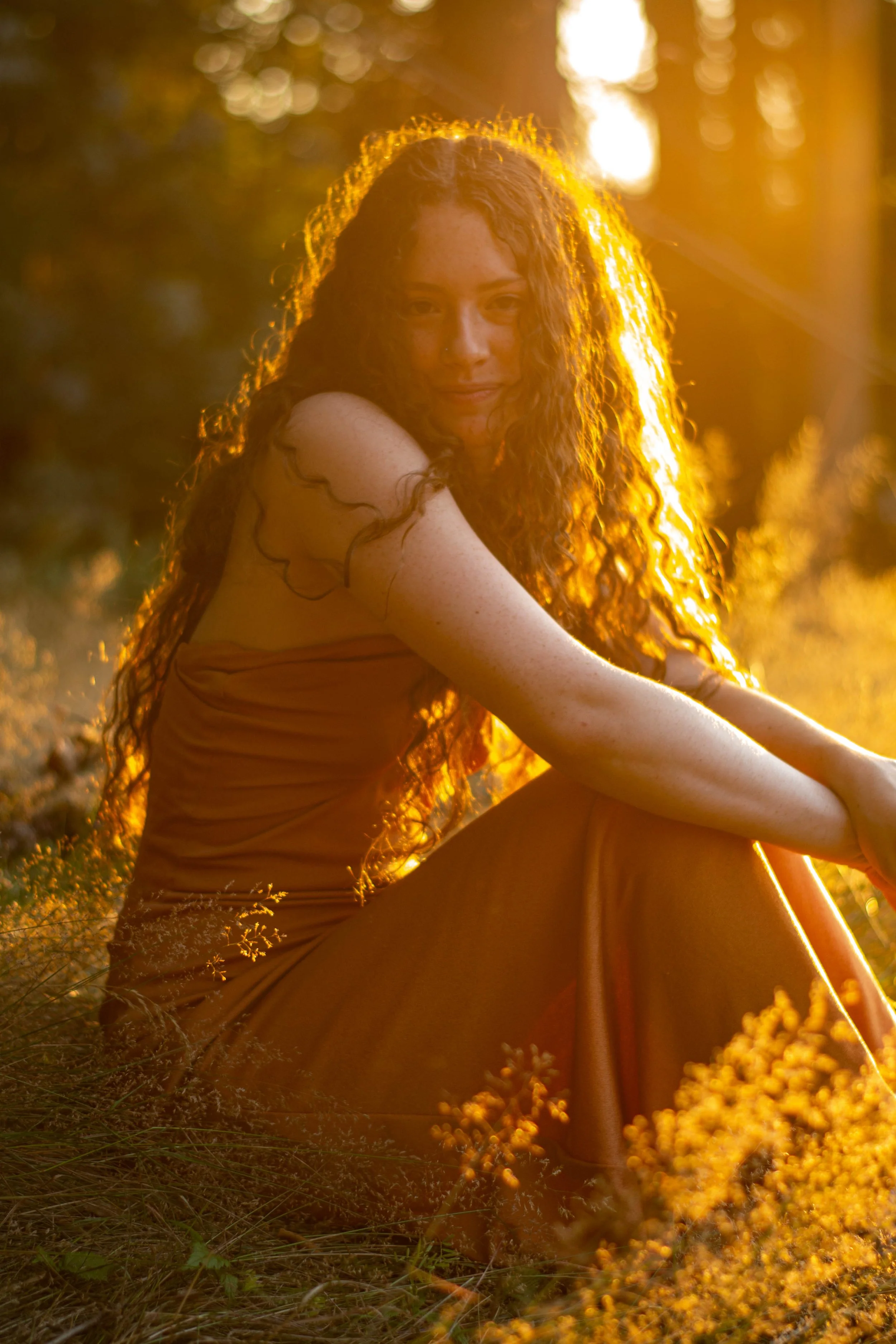 A woman with long, curly hair sitting outdoors during sunset, surrounded by tall grass and illuminated by warm golden light.