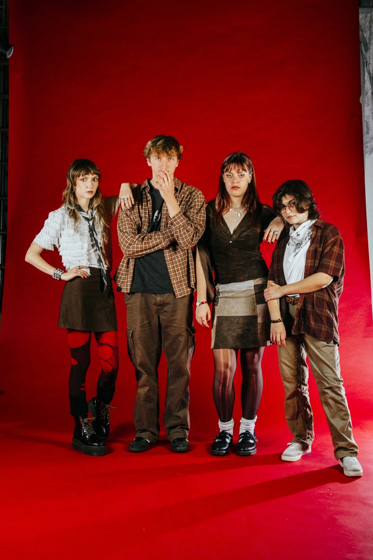 Four young adults standing together in front of a red backdrop. The group includes two women and two men, dressed in casual and grunge-inspired clothing with expressive hairstyles. They have a rebellious, alternative fashion style.