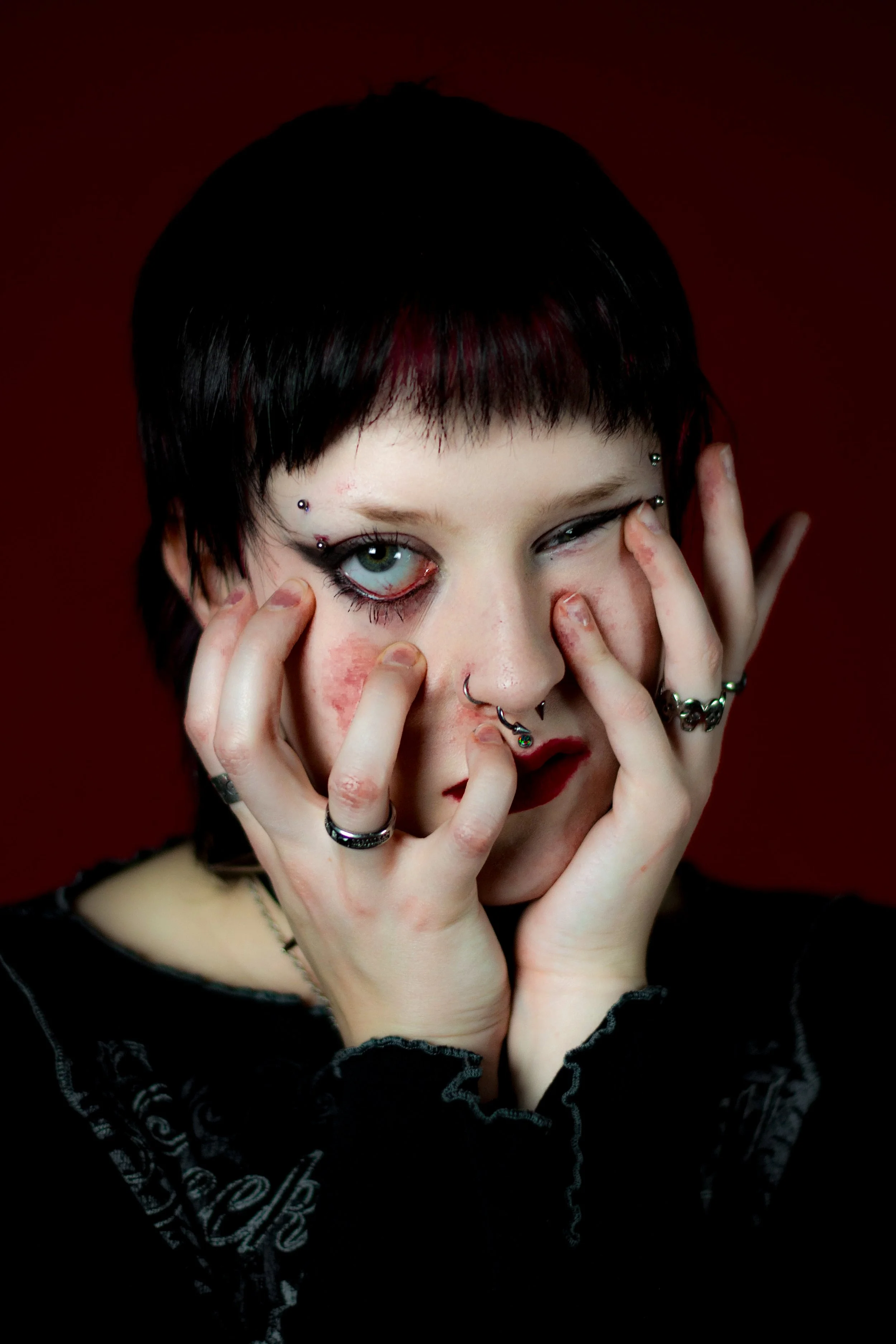 Person with short black hair and multiple facial piercings, including on eyebrows and lips. They have green eyes, dark makeup, and are touching their face with both hands, which are adorned with rings. The background is a dark red.
