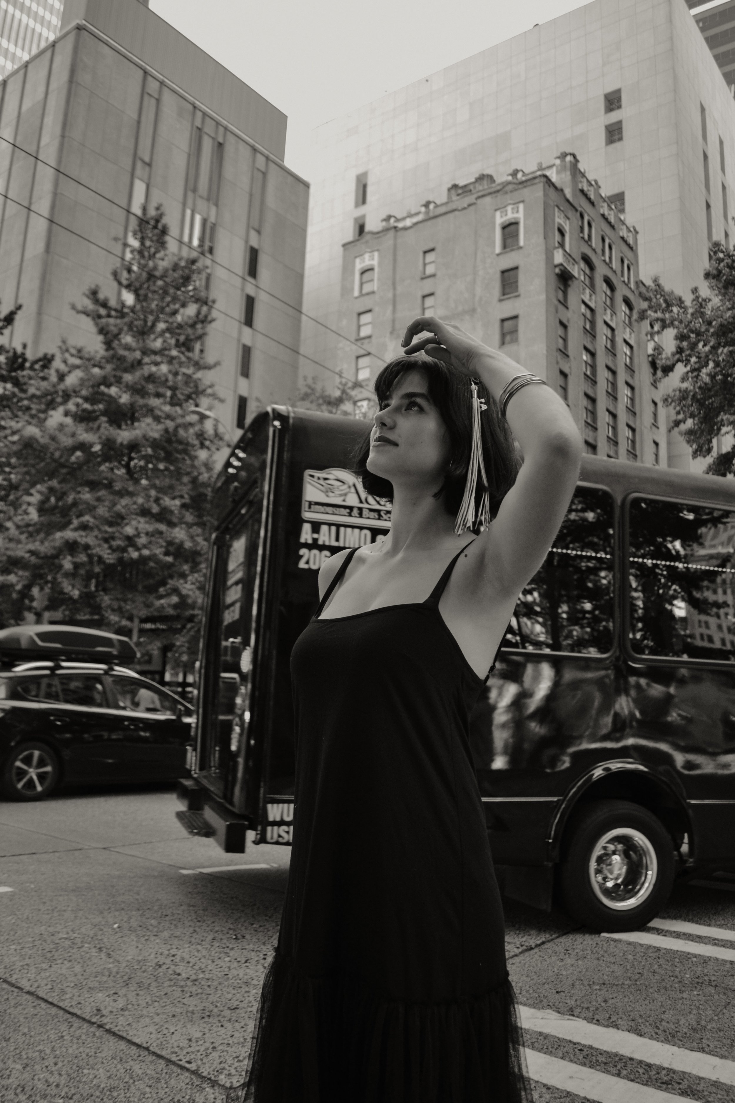 A woman standing on a city street, wearing a black dress and a tassel earring with her hand raised above her head, with tall buildings and trees in the background.