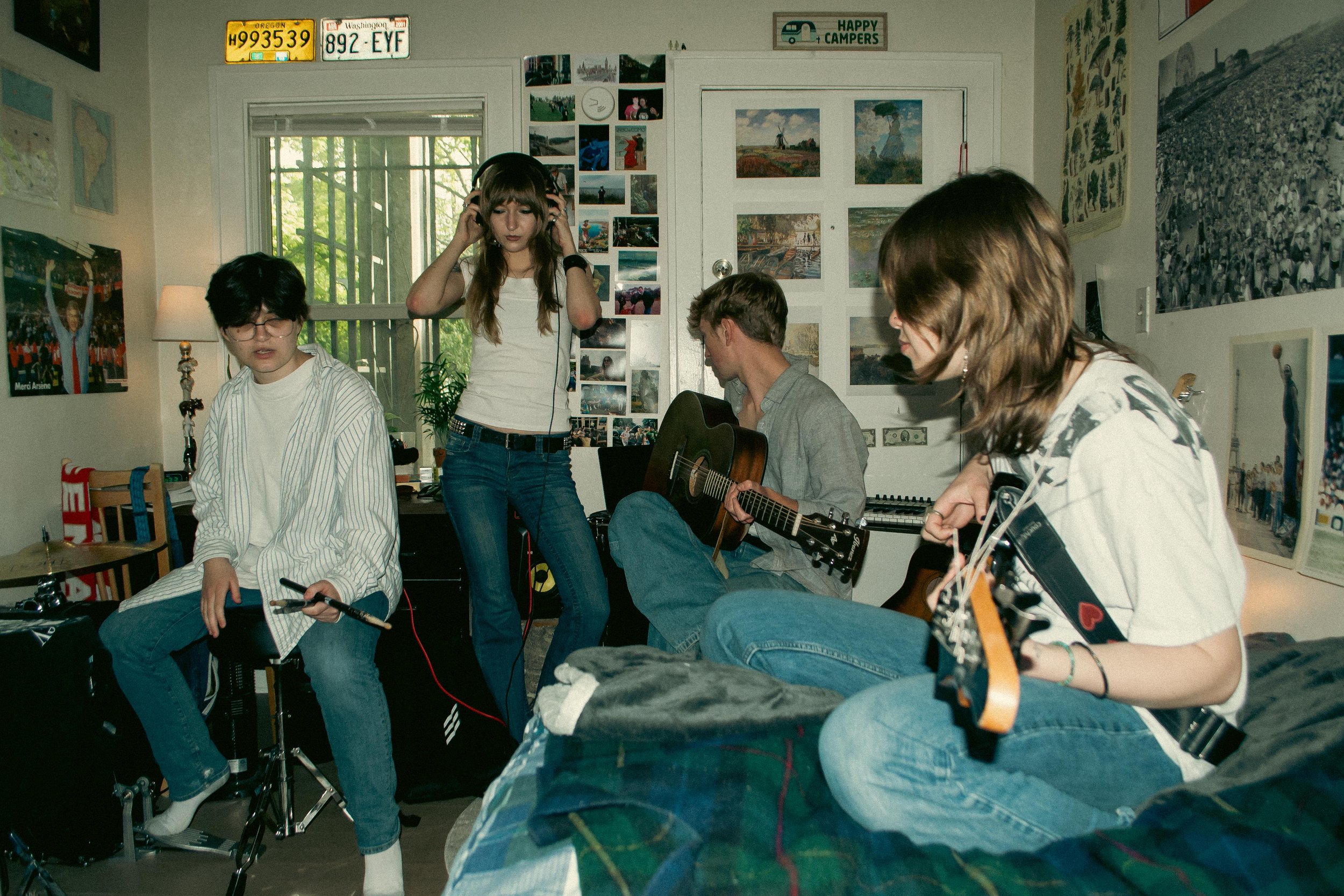 A group of young people in a room, some playing guitars and others listening, with posters and photos on the walls and license plates and signs above the window.