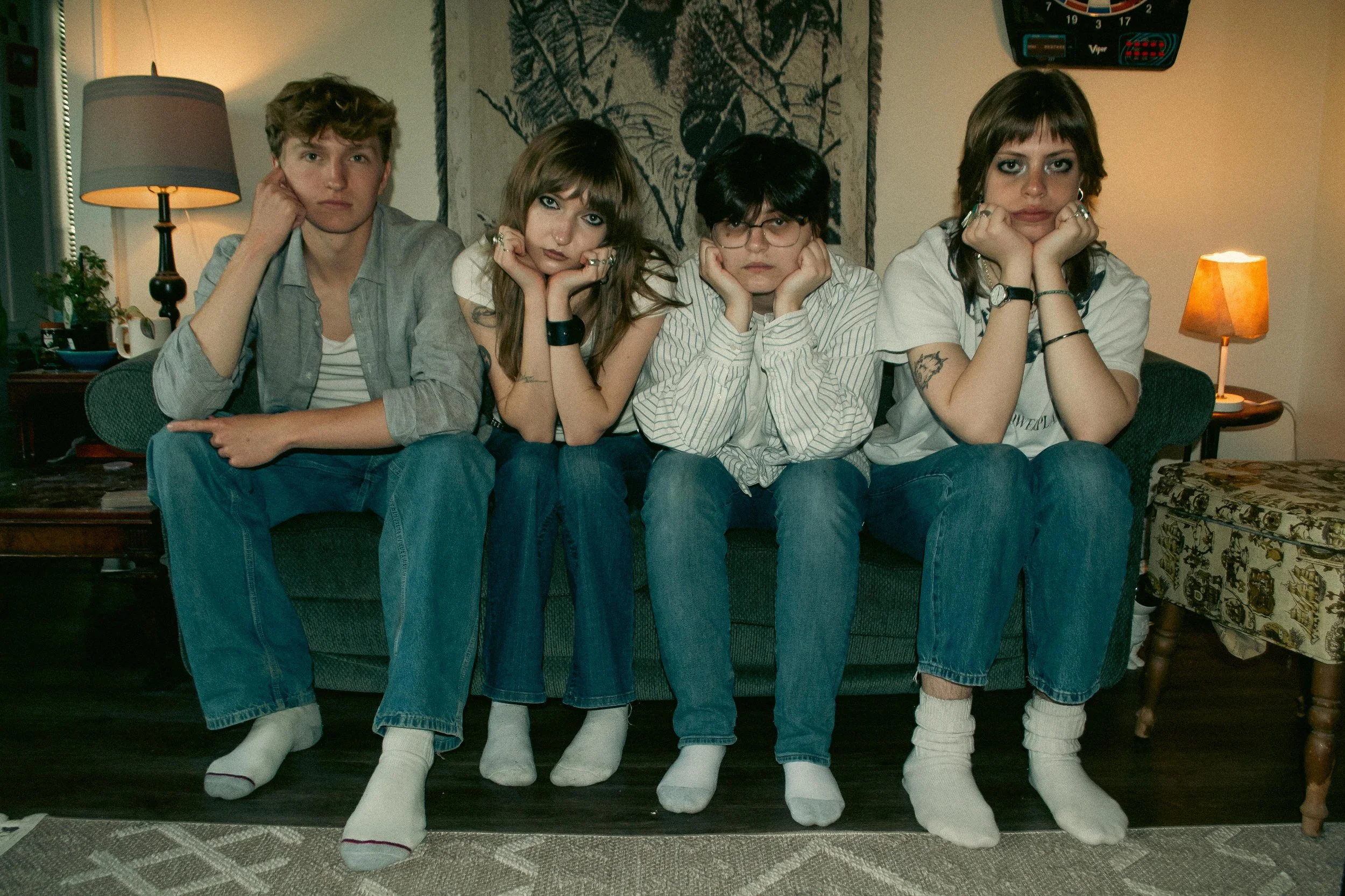 Four young adults sitting on a couch with their chins resting on their hands, looking directly at the camera. They are wearing casual clothes and socks, and have a relaxed, slightly bored expression. There are two women and two men, and the backgroun
