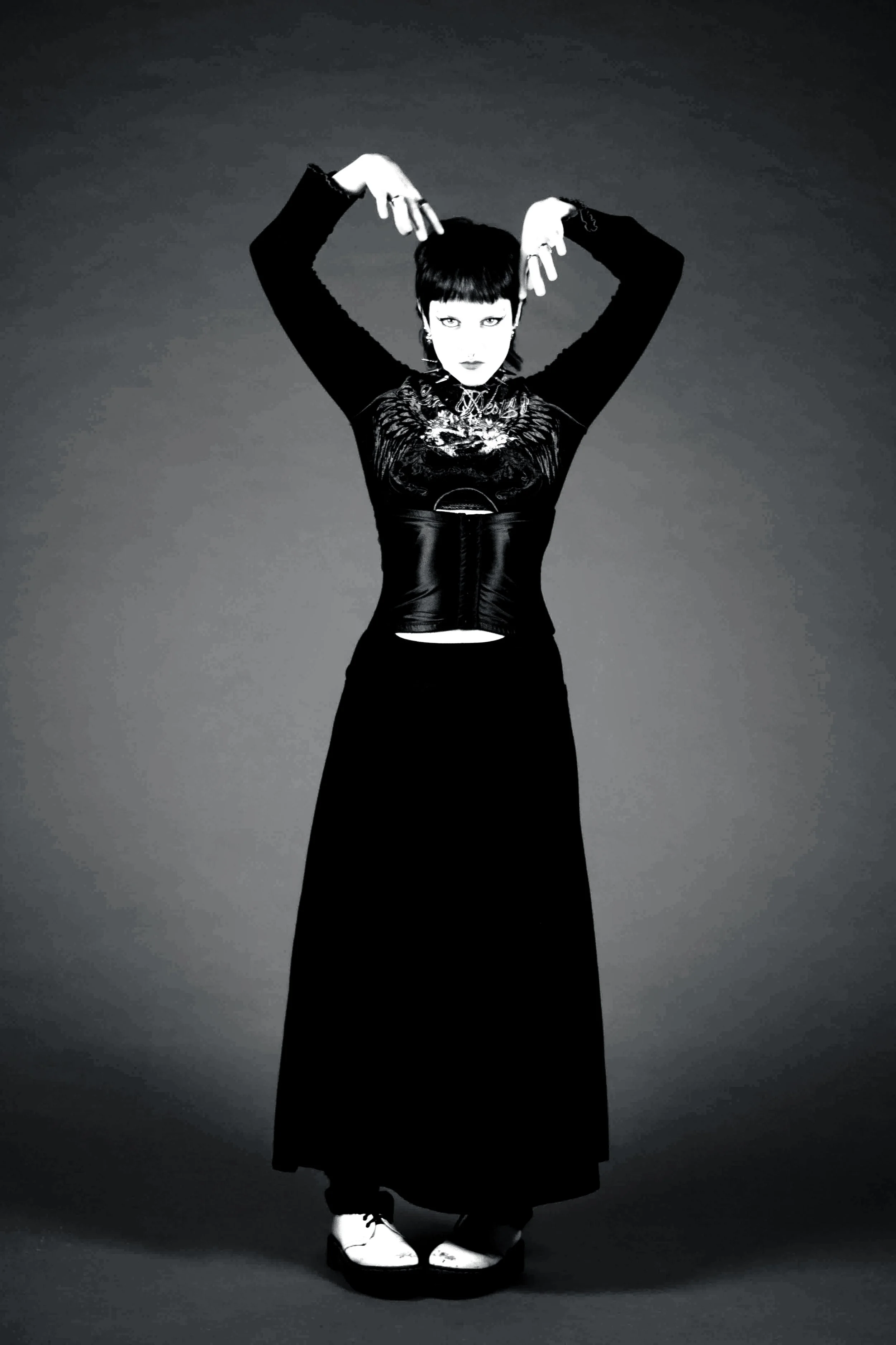 Black and white photo of a person with short hair, dressed in a long skirt and a top with embroidery, standing against a plain background. They are looking directly at the camera with a serious expression, with arms raised above their head and hands 