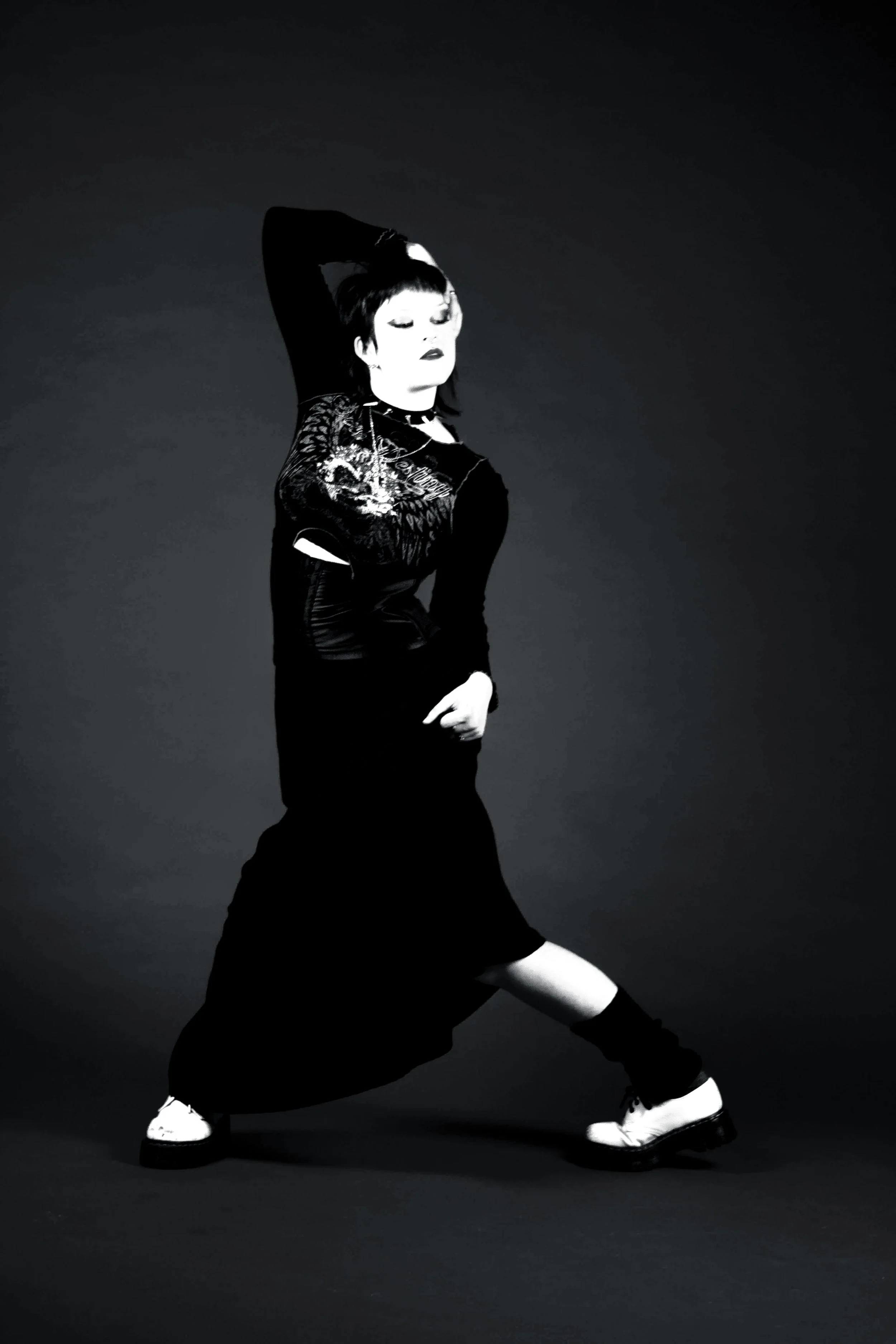 Black and white photo of a woman dancing or posing with one arm over her head and wearing a long skirt and sneakers, smiling with eyes closed.