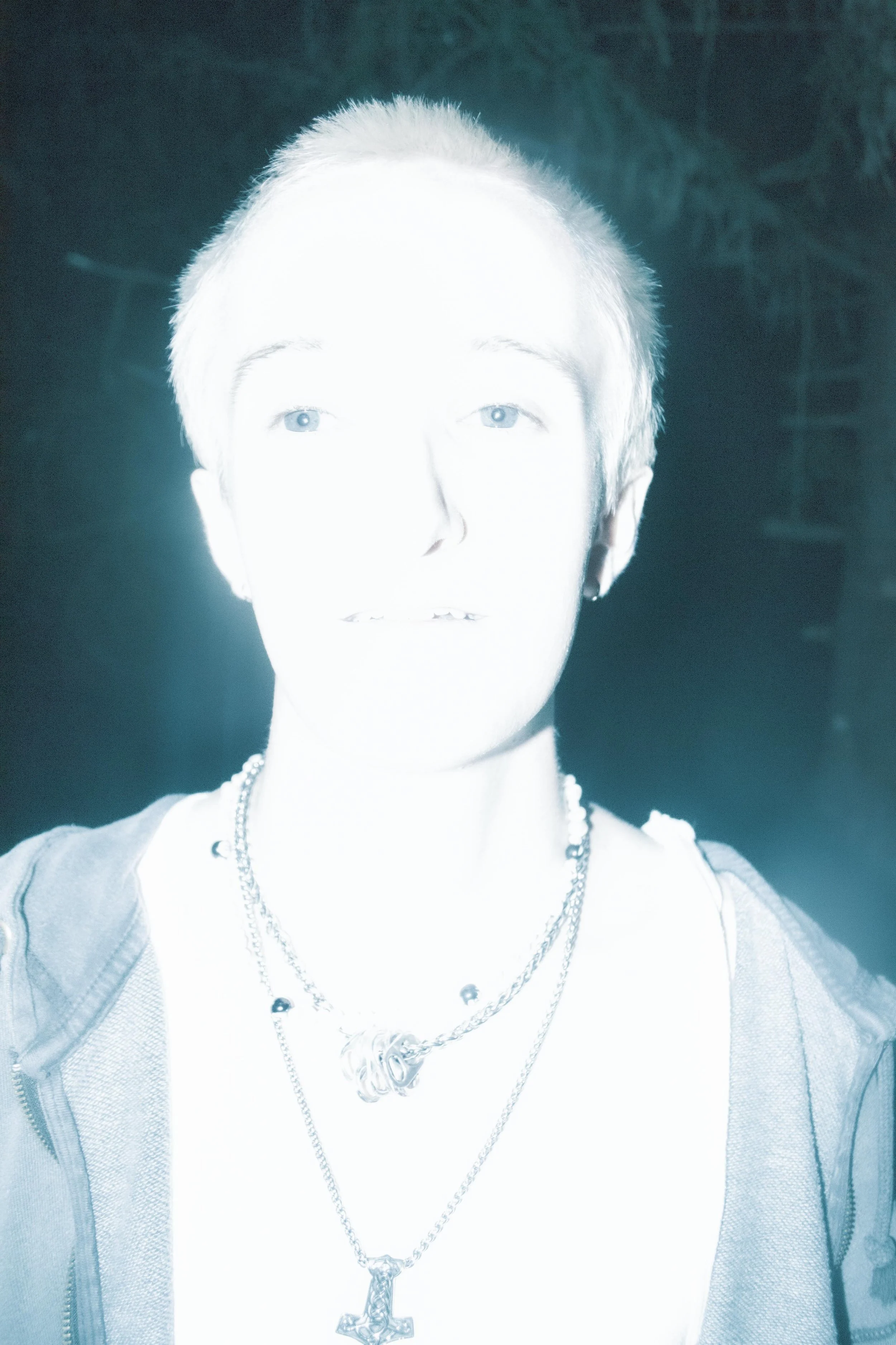 A person with short, spiky blonde hair wearing layered necklaces, a white shirt, and a gray hoodie, standing outdoors at night.
