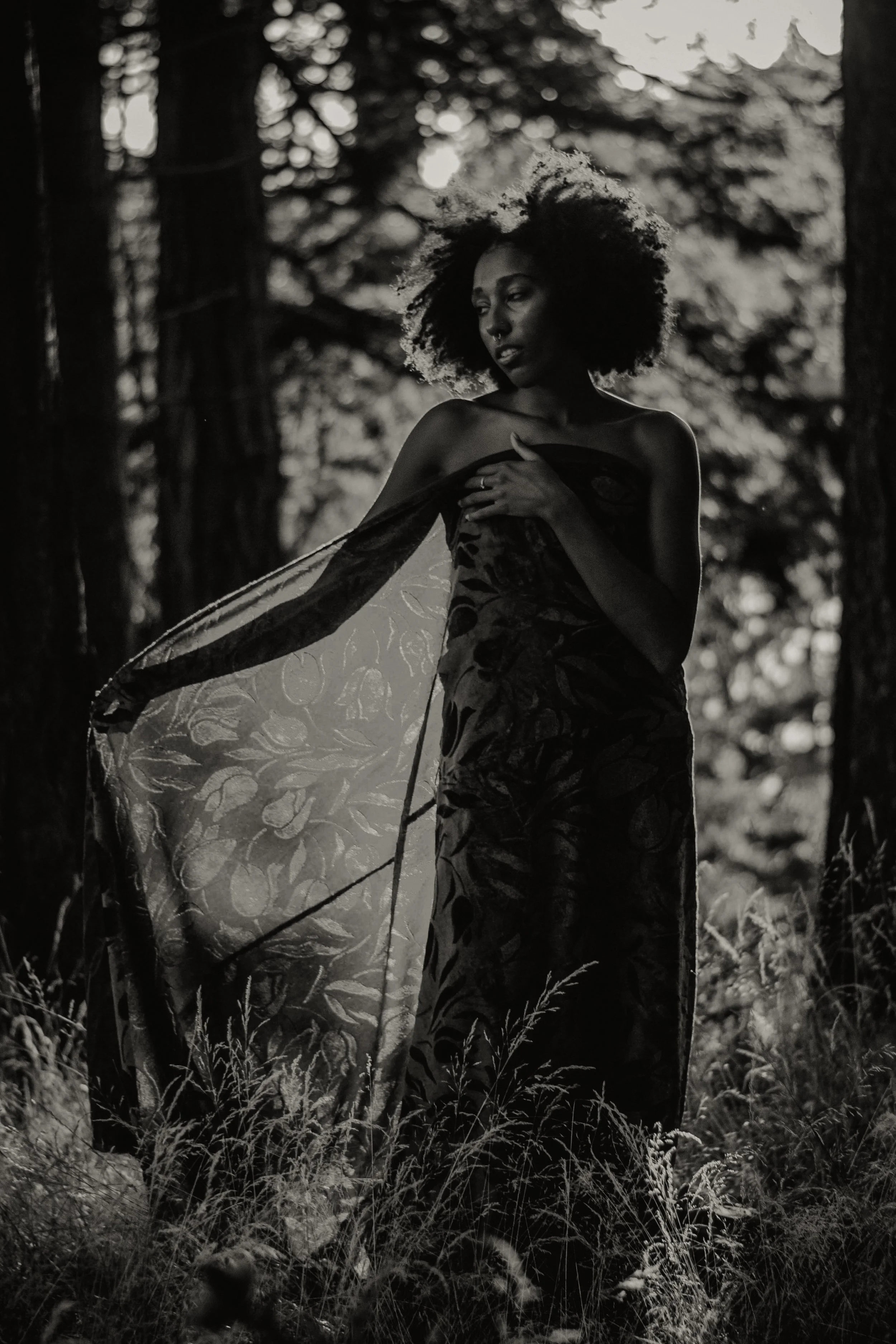 A woman with natural curly hair standing in a forest, wearing a patterned dress and a sheer fabric draped over her shoulder, with her hand on her chest, in black and white photography.