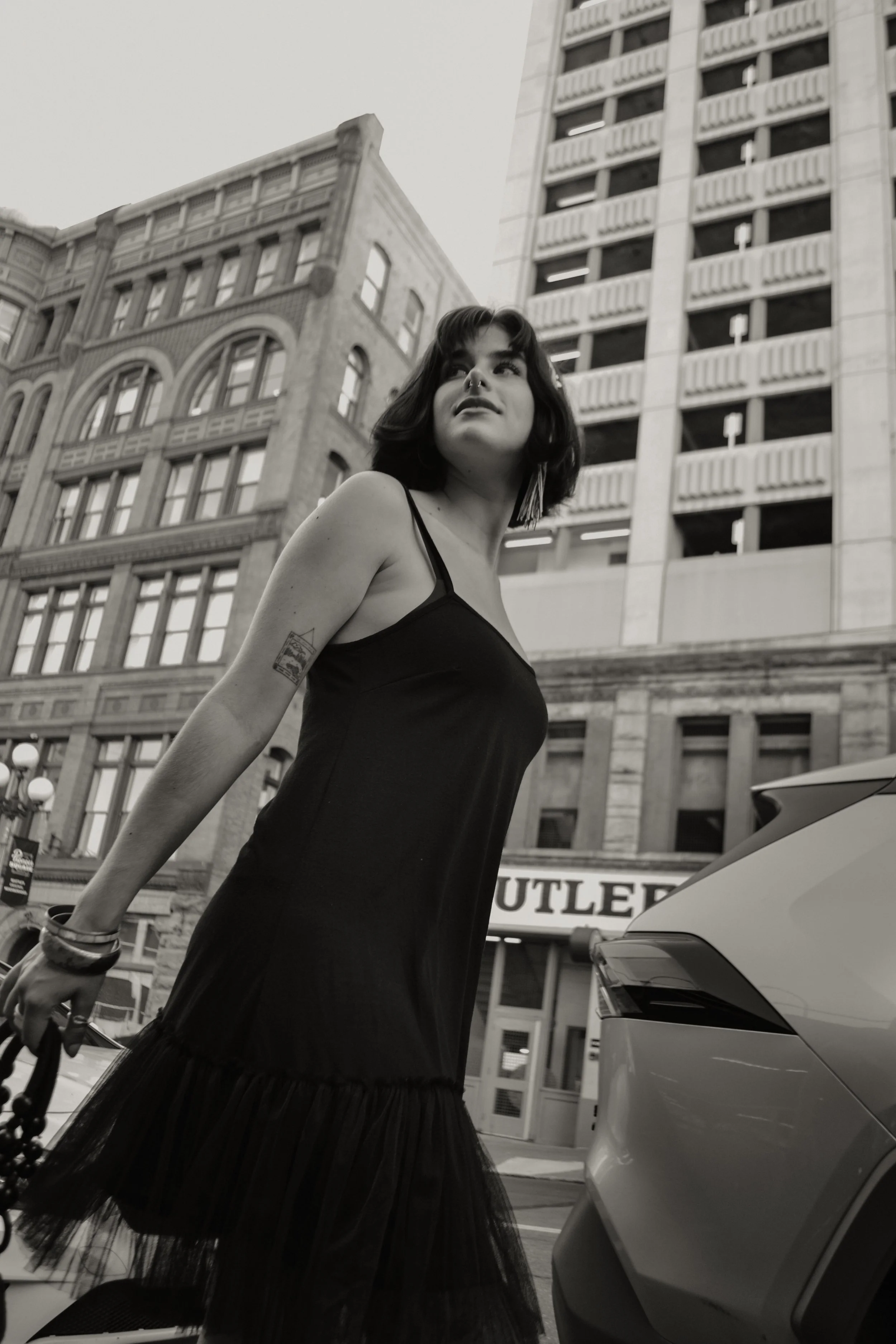 A woman with dark hair, wearing a black dress, stands outdoors near cars and buildings in an urban setting.