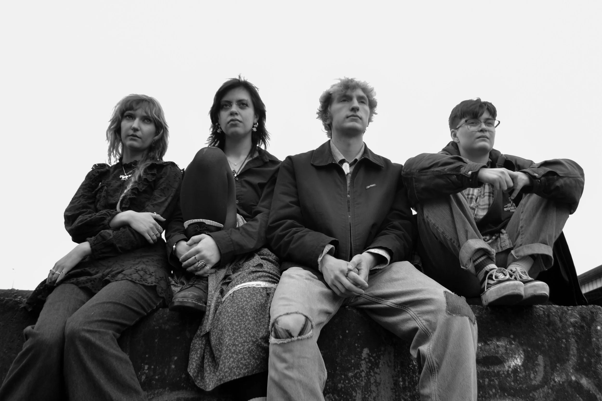 Black and white photo of four young people sitting outdoors on a large rock, looking serious and contemplative, with overcast sky in the background.