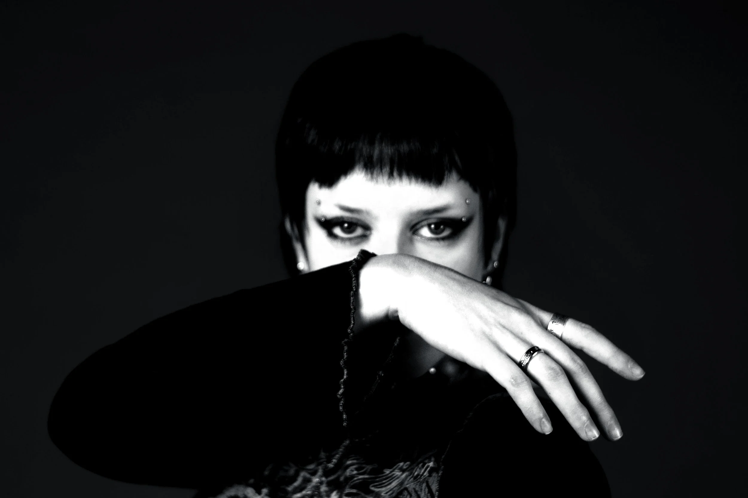 A black and white portrait of a person with short dark hair and intense eyes, covering part of their face with their hand. They are wearing rings and earrings.