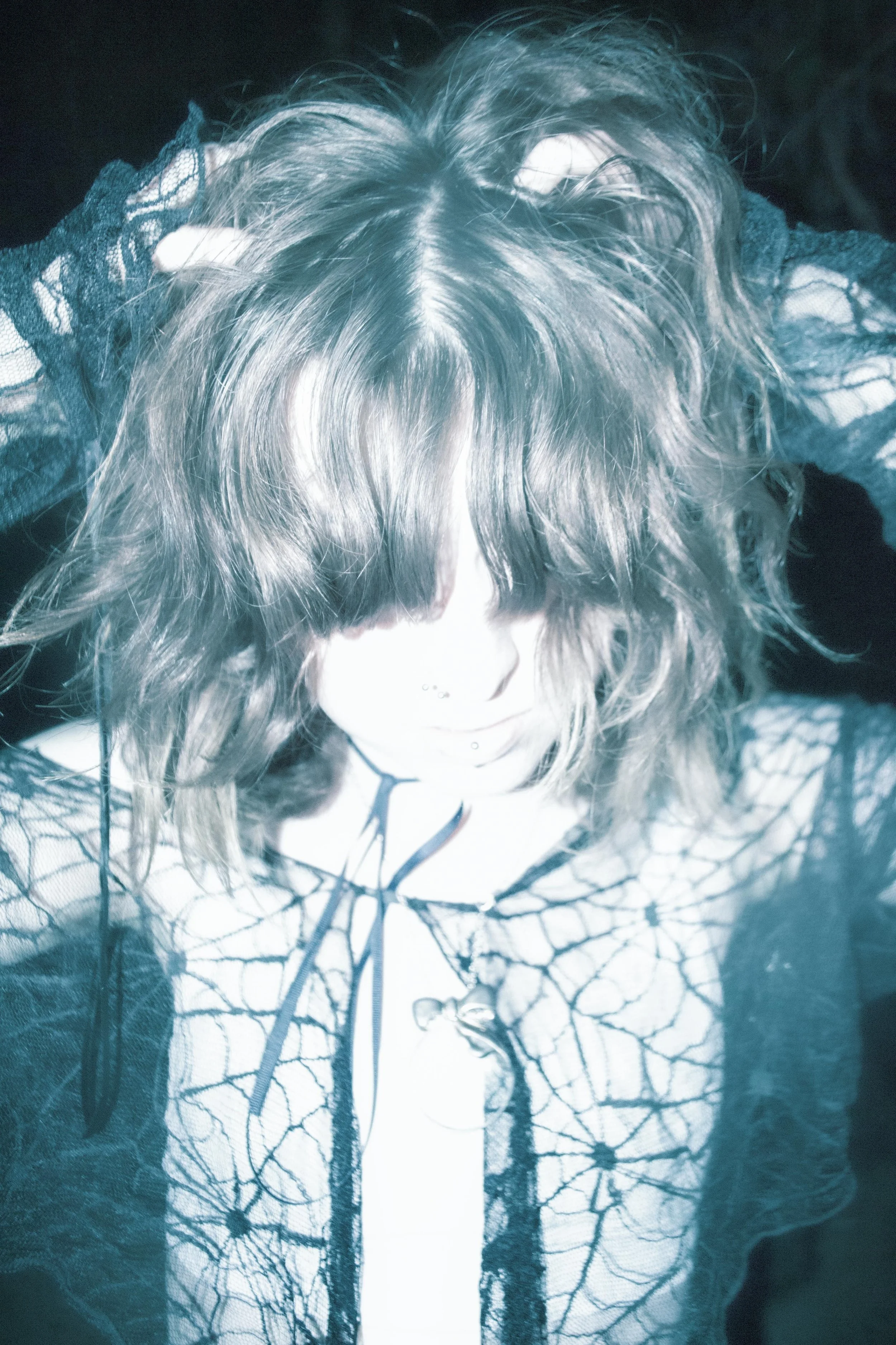 A person with shoulder-length curly hair is looking down, wearing a black lace top and a jacket featuring a tree branch pattern. The image has a cool, bluish tint.