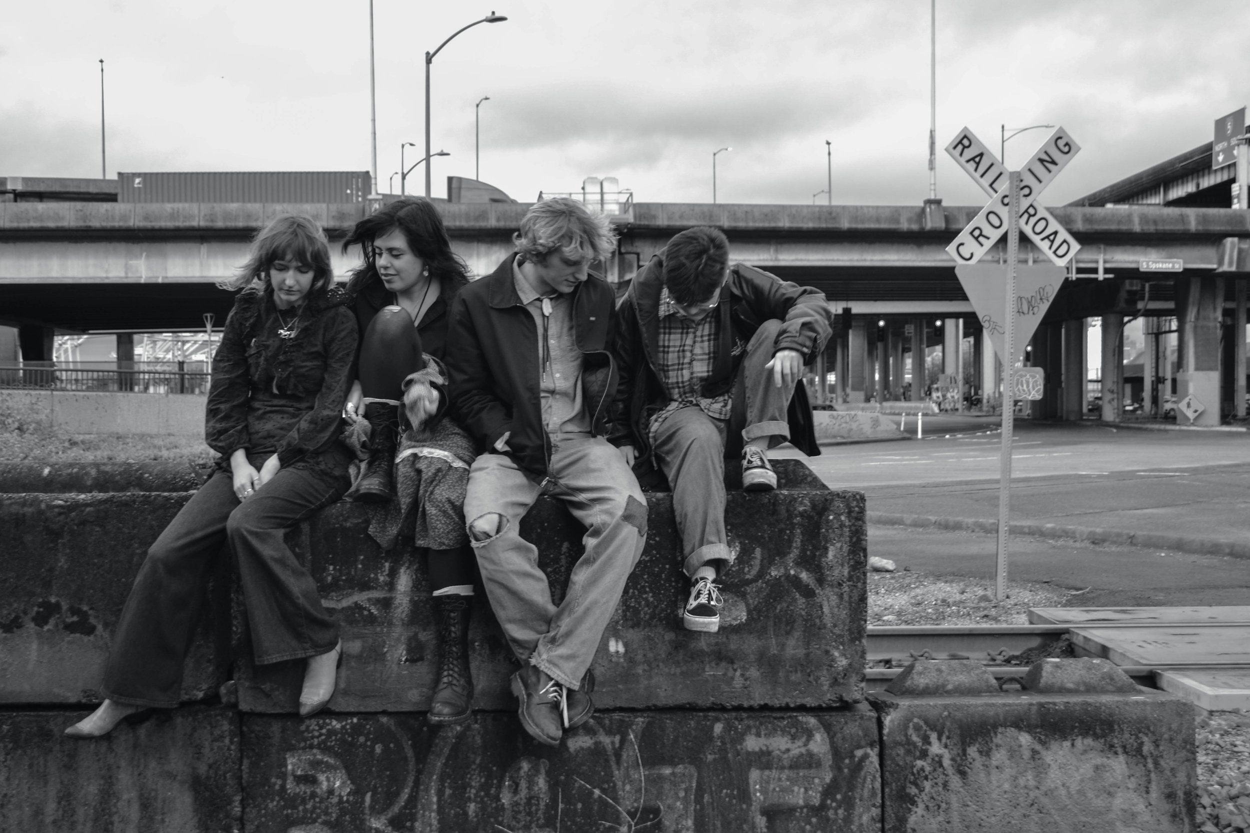 Four young people sitting on a concrete ledge at a train crossing, with a railroad crossing sign and overpass in the background. The image is in black and white.