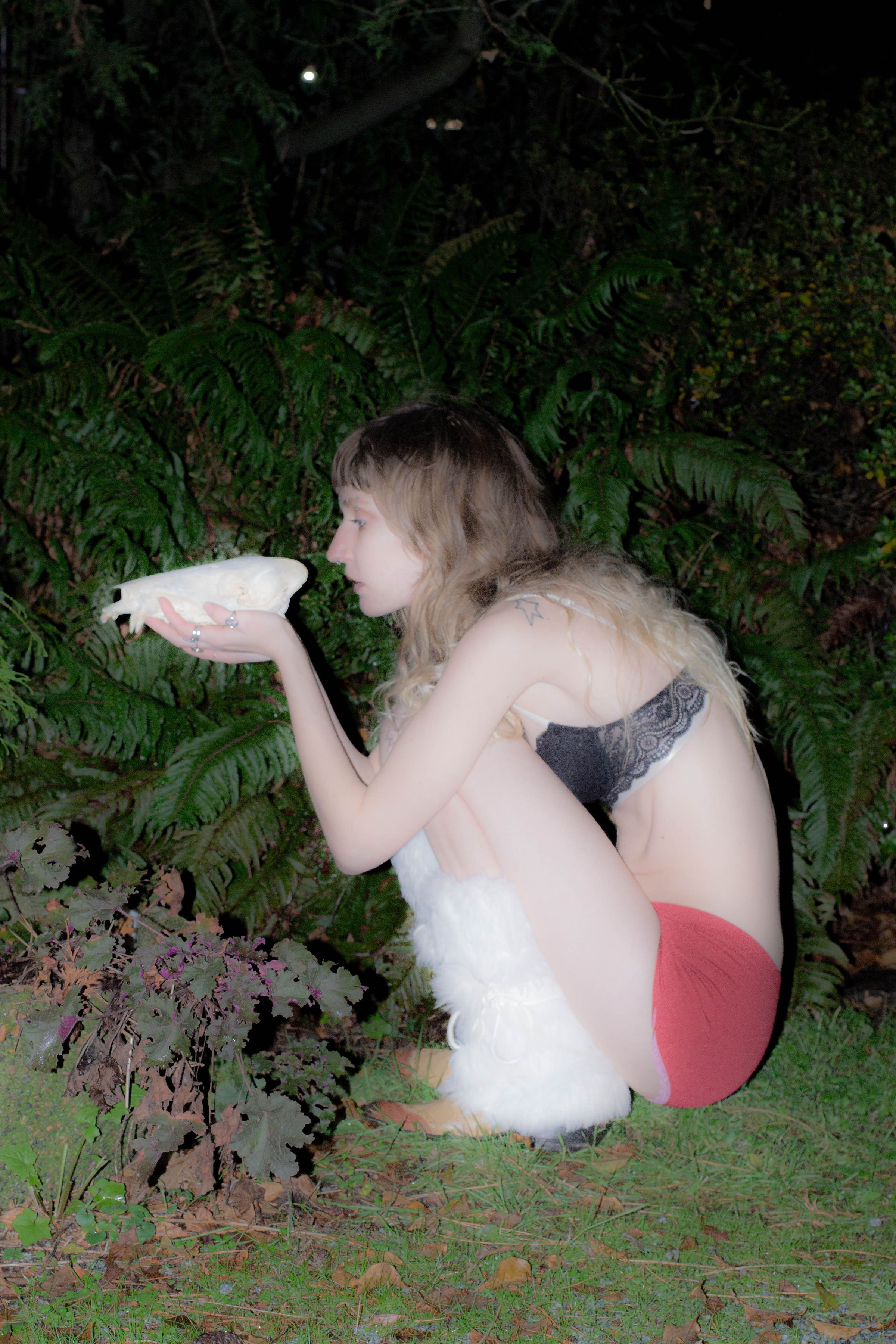 Young woman with light brown hair and light skin, wearing red shorts and a black lace bra, crouching outdoors among greenery, holding a carved white skull in her hands.