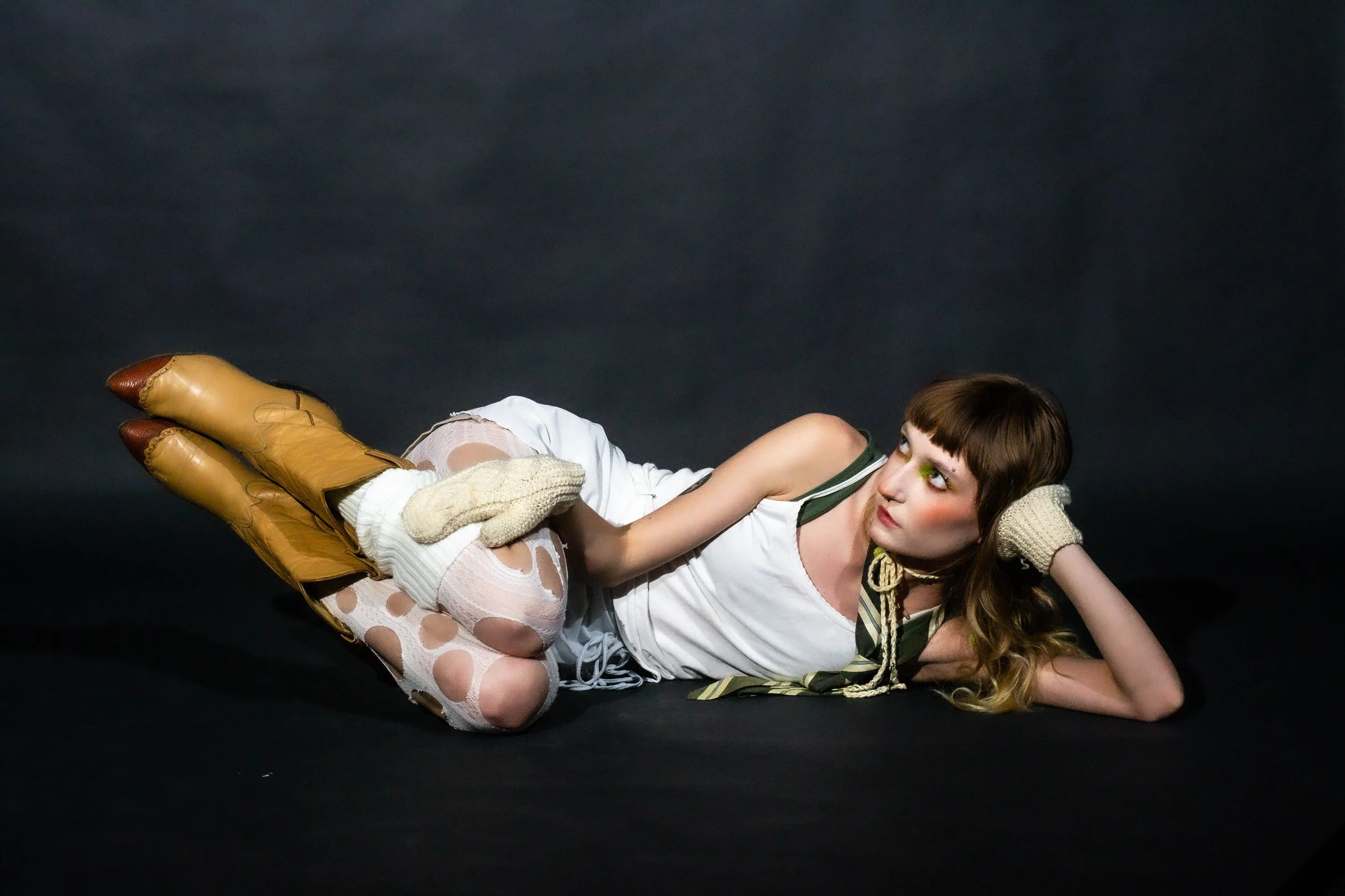 A woman lying on her side on a black background, wearing a white shirt, ripped fishnet stockings, and tan boots, with her head resting on her hand and looking towards the camera.