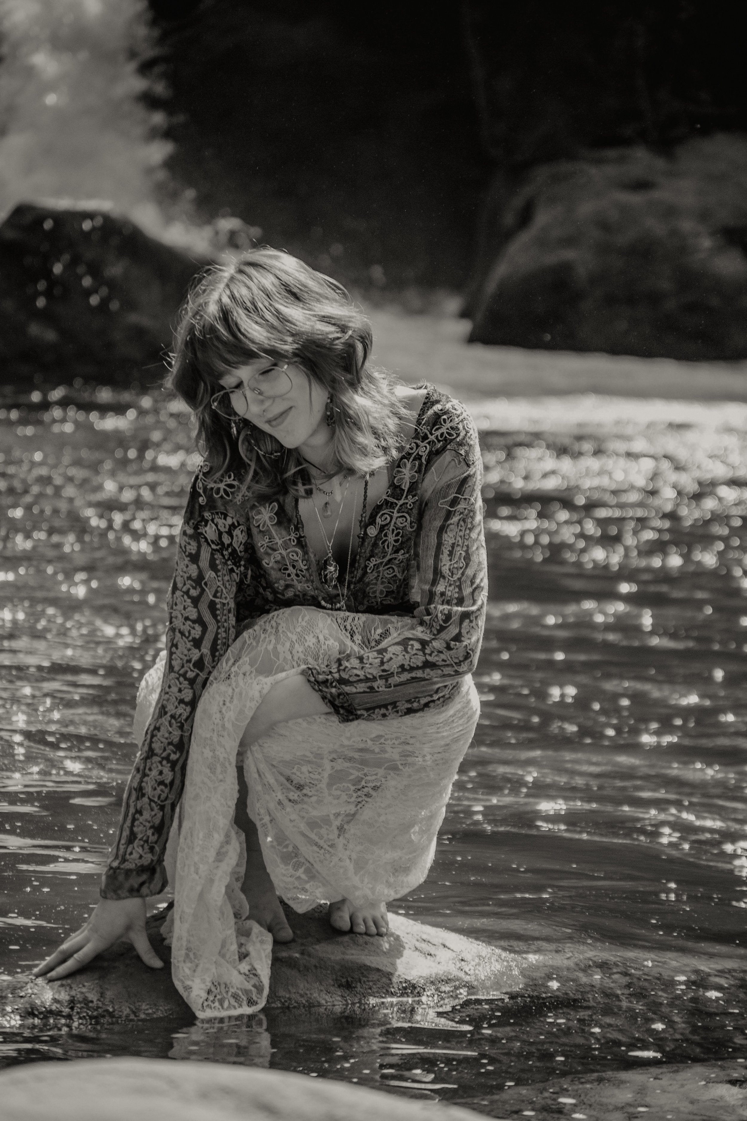 A woman with glasses and wavy hair kneels in shallow water at the beach, smiling softly, wearing a patterned long-sleeve blouse and lace skirt, with rocks and water in the background.