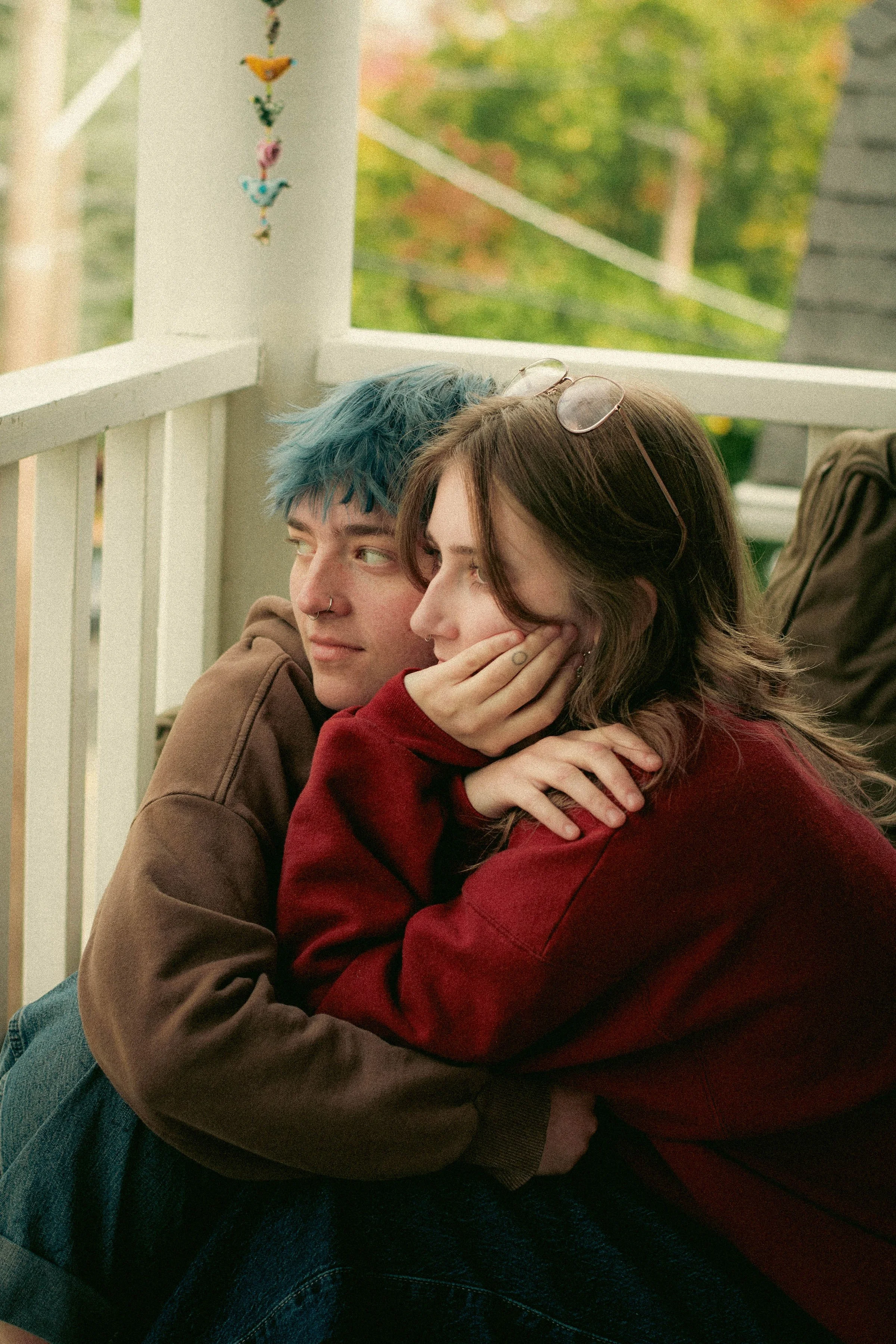 Two friends sitting closely on a porch, hugging and leaning on each other, with colorful autumn trees in the background.