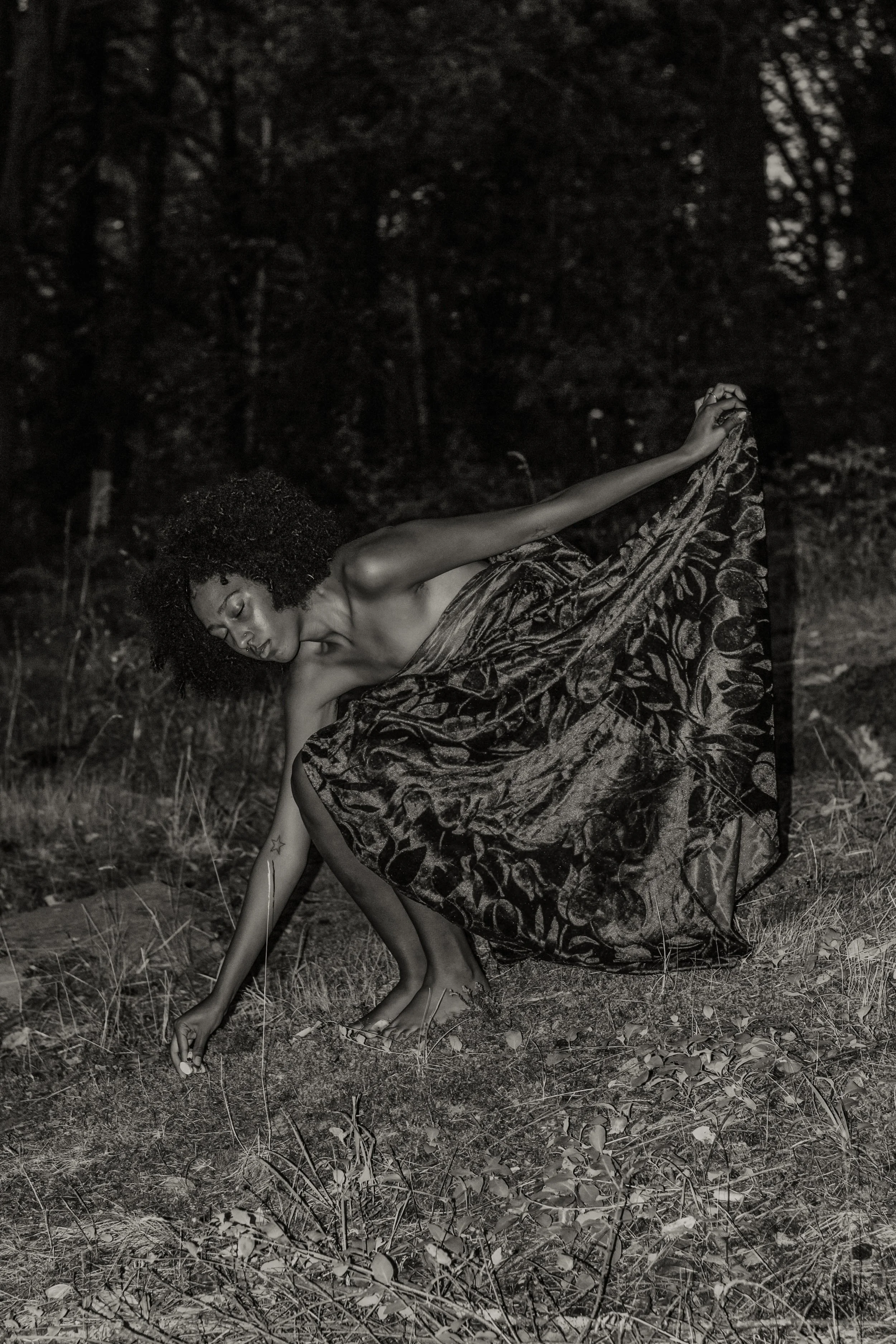 A woman with curly hair in a patterned dress bending down outdoors at night, surrounded by trees and grass.