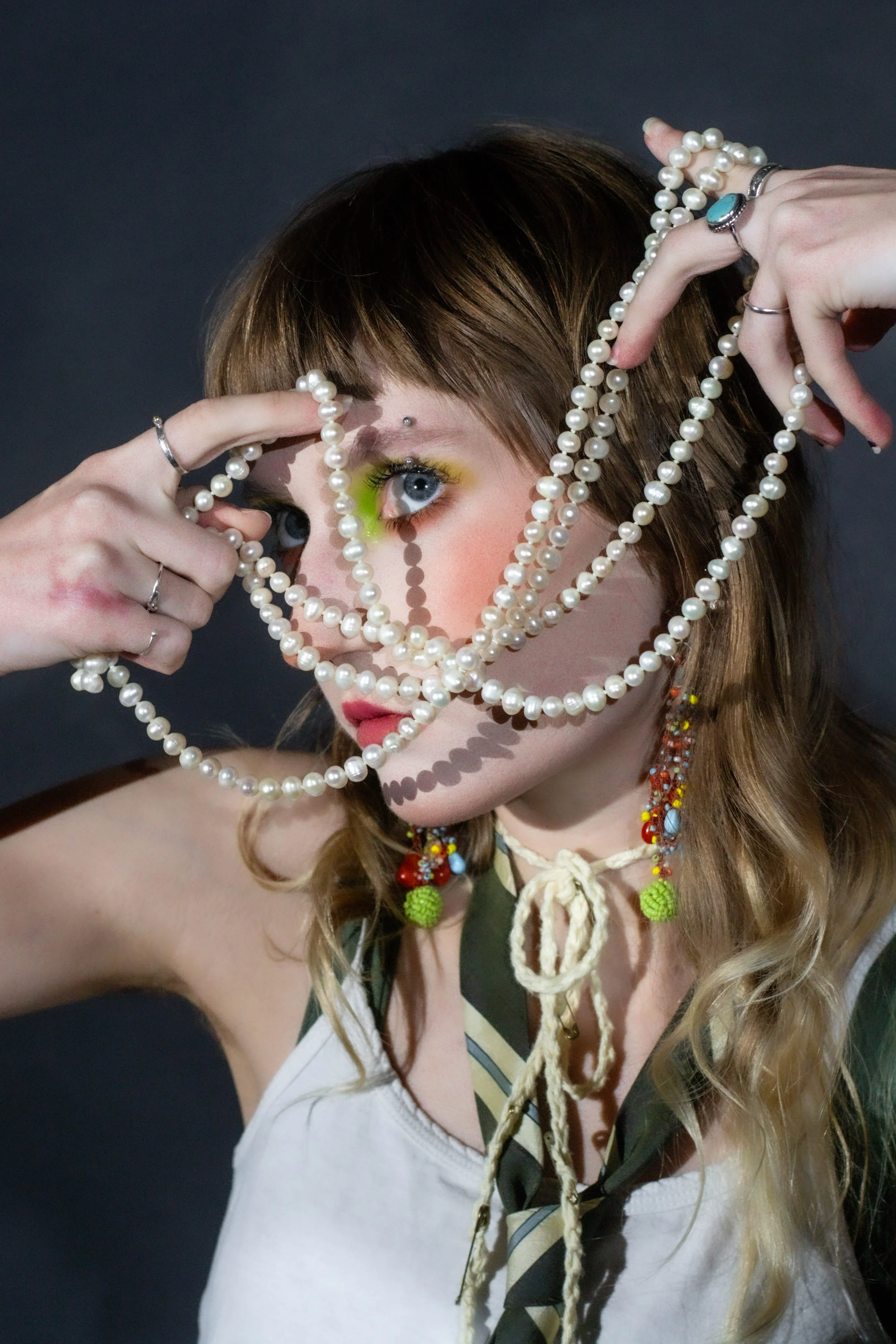 A woman with light skin and brown hair styled in loose waves, wearing a white top. She has bright makeup with yellow eyeshadow and a silver eyebrow piercing. She is holding a long pearl necklace in front of her face, partially covering her eyes, and 