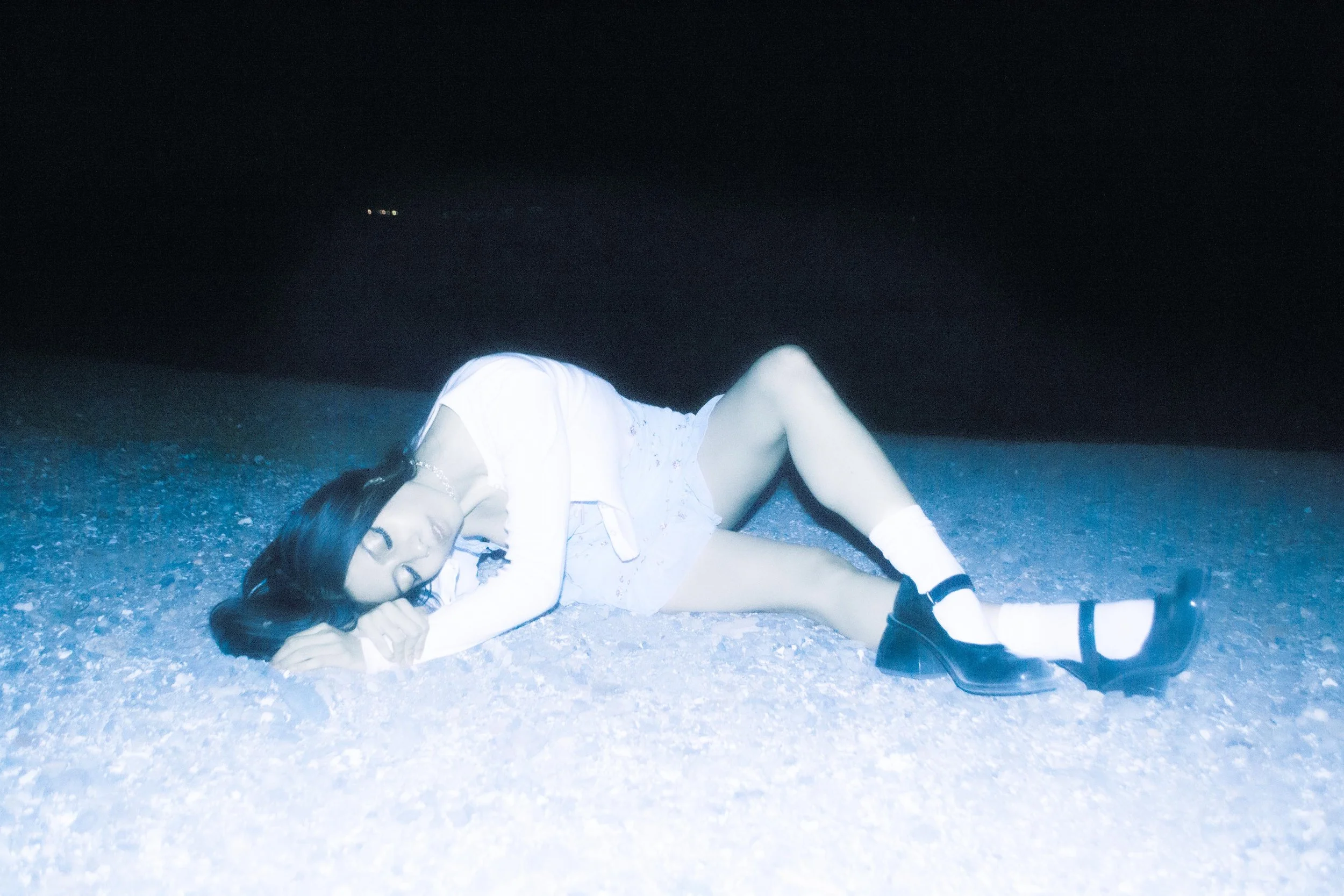 A woman lying on a gravel surface at night, wearing a white dress with lace details, black boots, and socks, with her eyes closed and long dark hair spread out around her head.