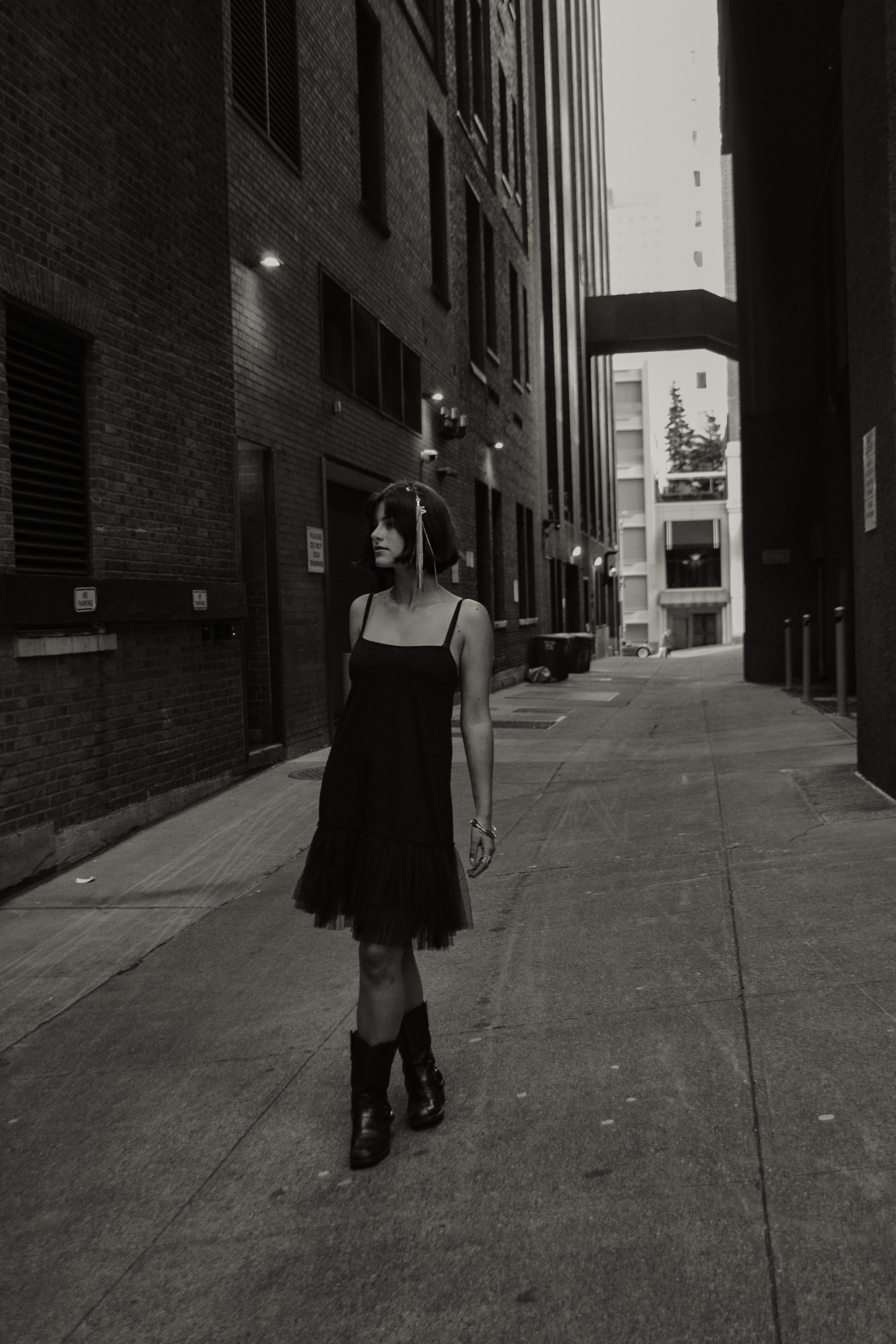 A woman in a black dress and combat boots walking through a narrow urban alleyway, with brick and concrete buildings on either side, and a distant view of a car and some trees.