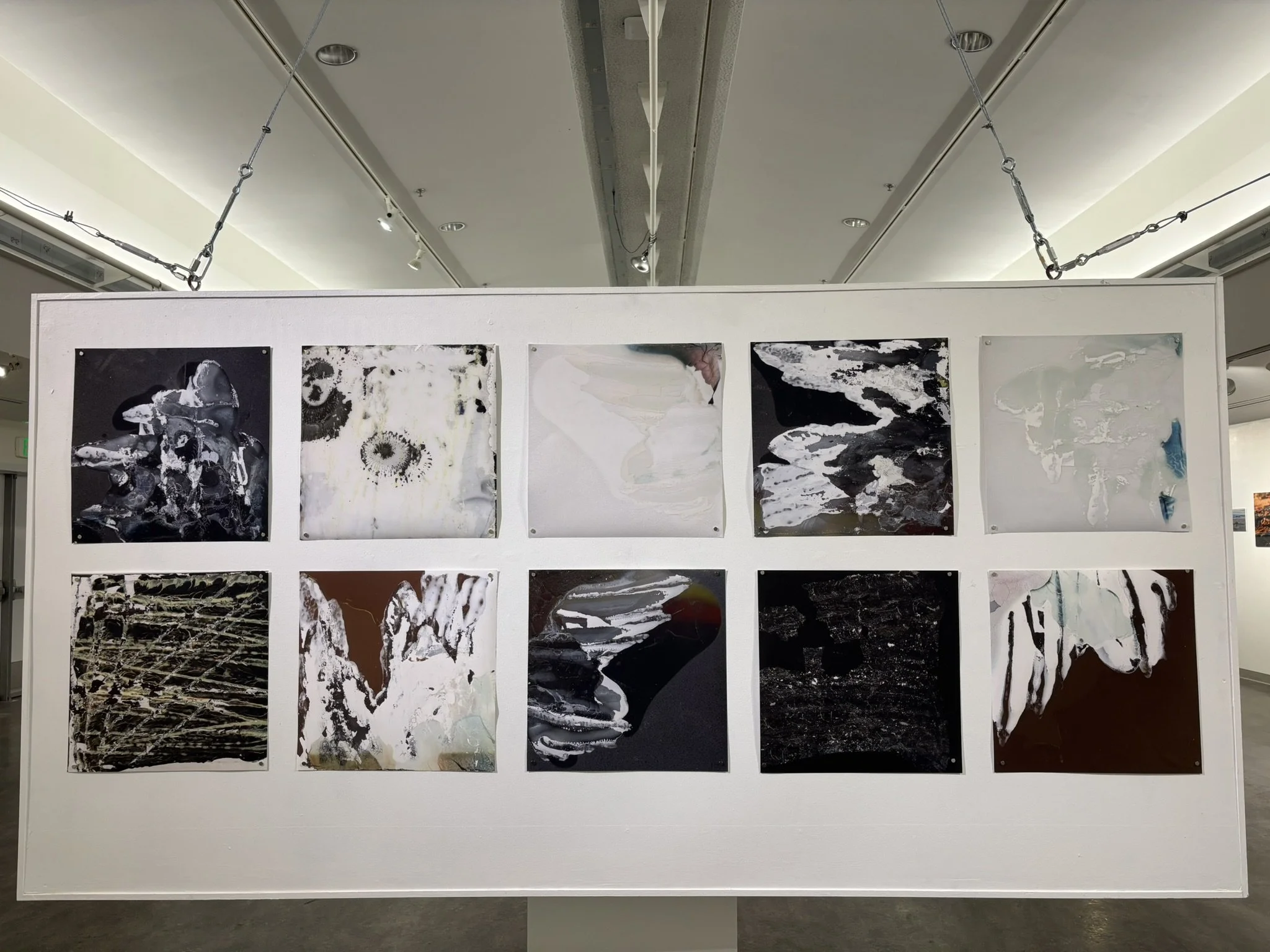 An art exhibit featuring abstract paintings displayed on a white wall in a gallery. The ceiling has track lighting and chains for hanging the artwork.