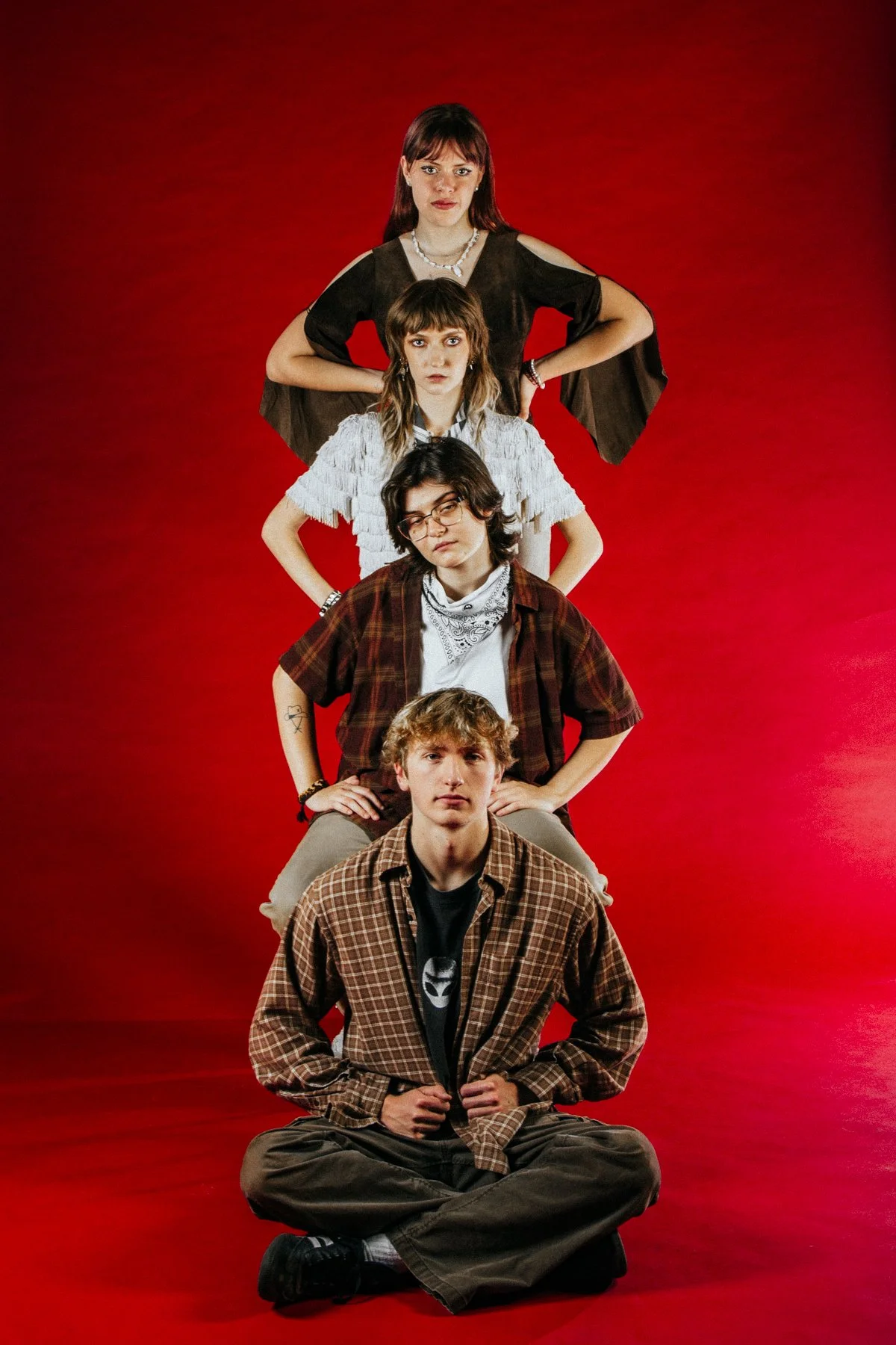 A group of five young people are stacked in a line, sitting on the floor against a red background. From bottom to top: a young man with curly hair and a plaid shirt, a young person with glasses and shoulder-length hair, a young woman with shoulder-le
