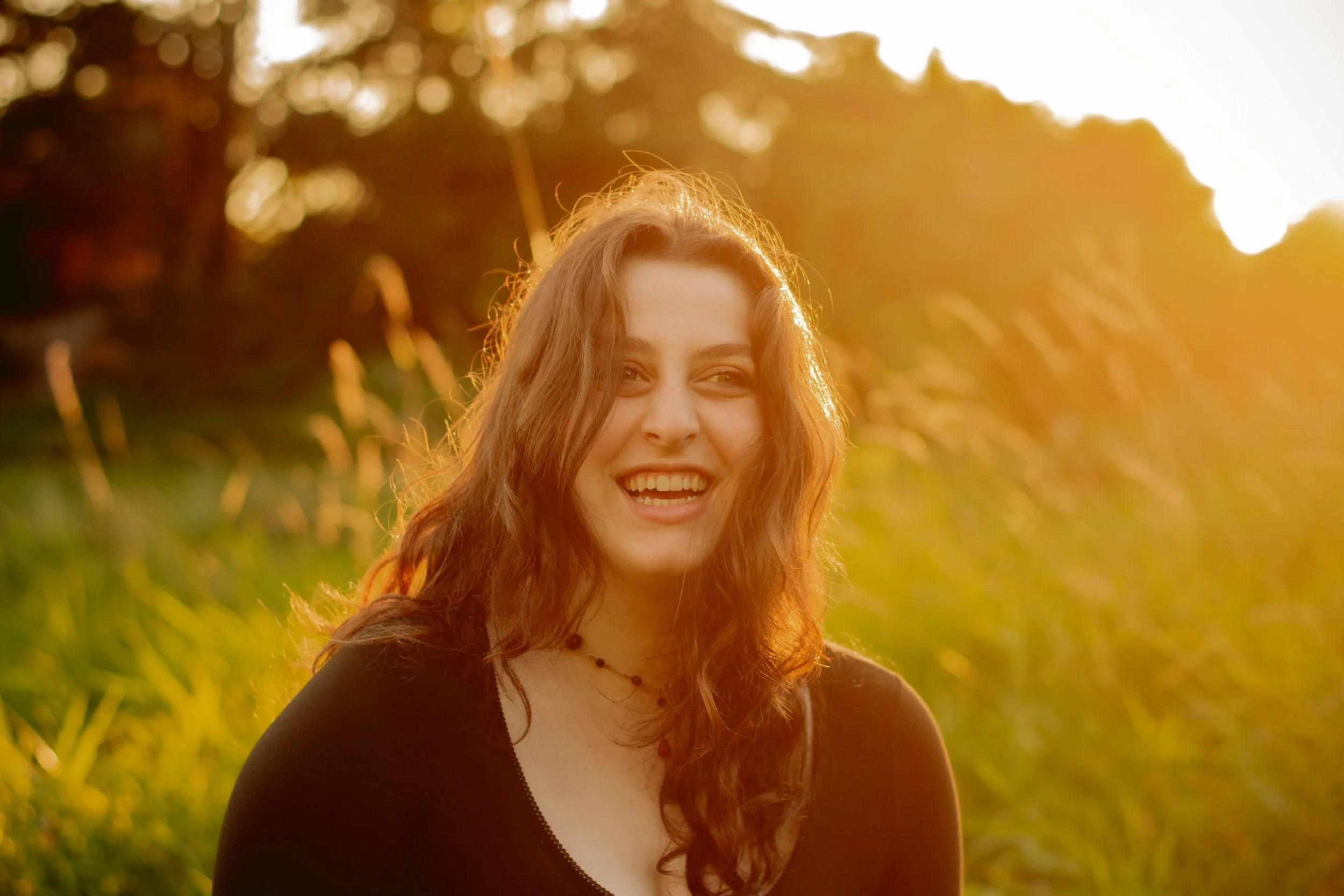 A woman with long curly reddish-brown hair smiling outdoors in a field during golden hour.