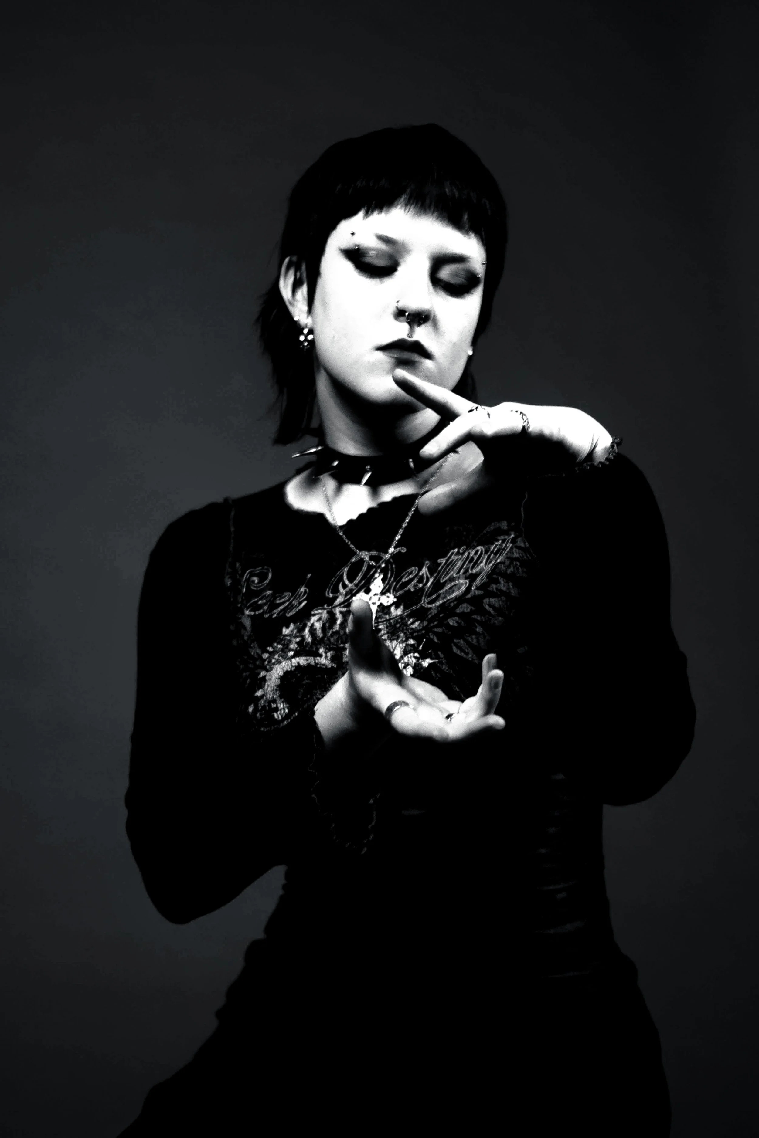 Black and white photo of a young person with dark hair and bangs, wearing multiple earrings and rings, making a gesture with their hands and looking down with a serious expression.