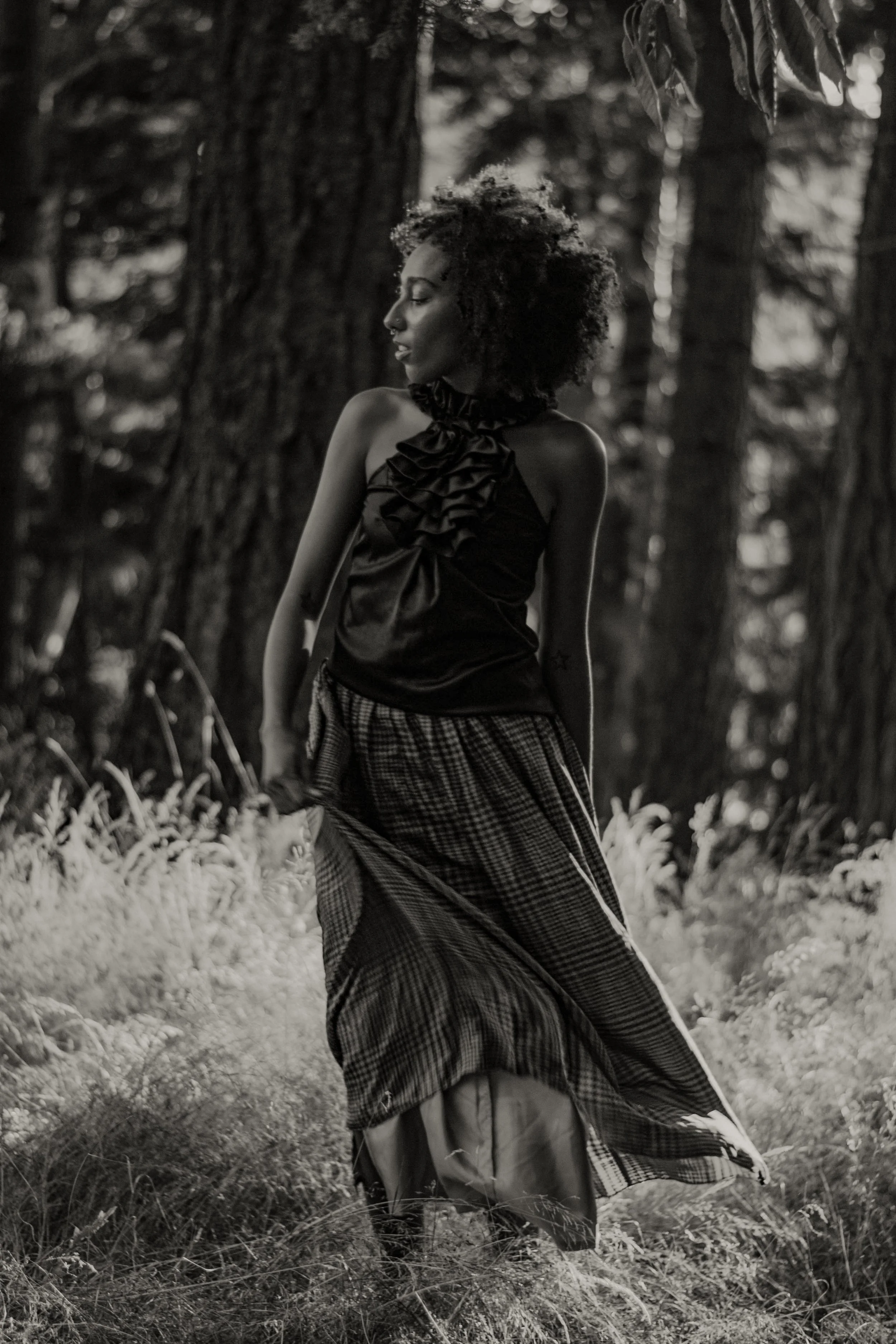 A young woman with curly hair, wearing a sleeveless top and a patterned skirt, stands outdoors in a forested area with tall trees and grassy ground. She has a neutral expression and is looking downward with her eyes closed.