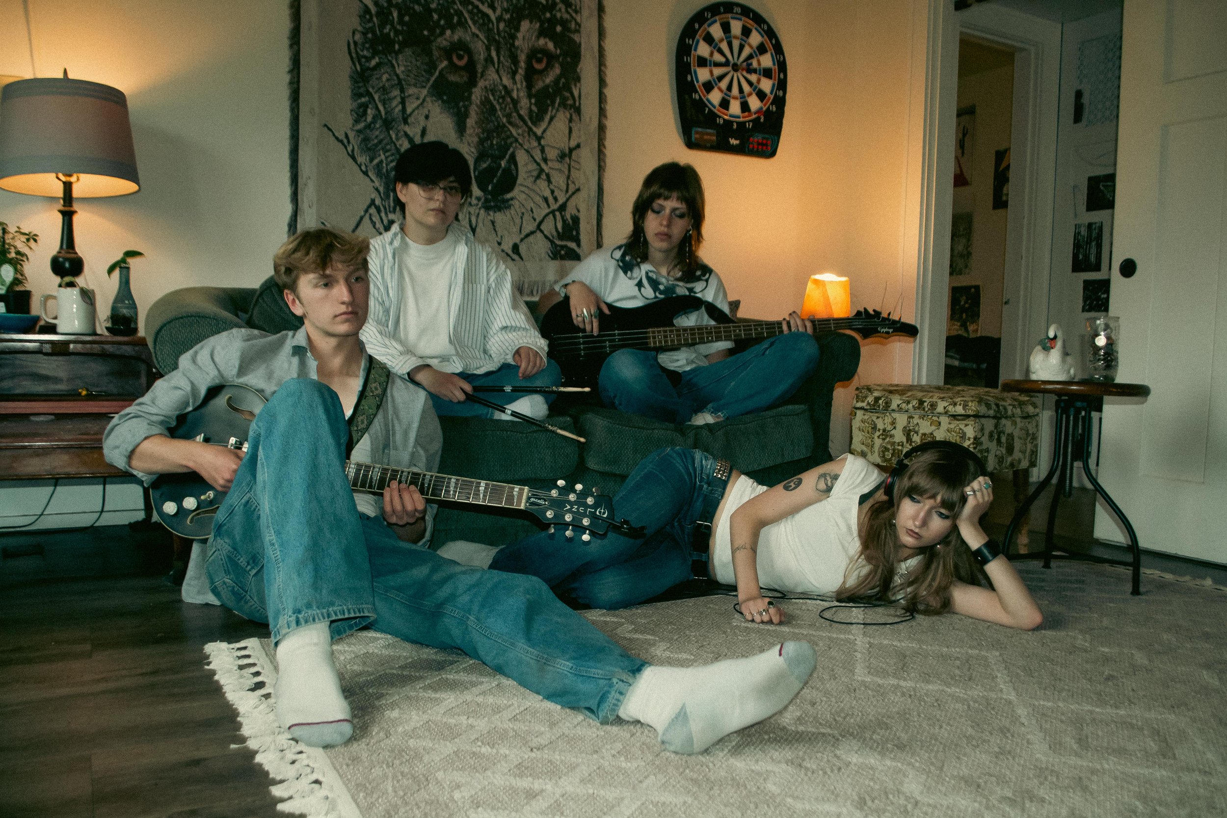 A group of five young people relaxing in a living room, with some playing musical instruments and others resting, with a dartboard and animal tapestry on the wall in the background.