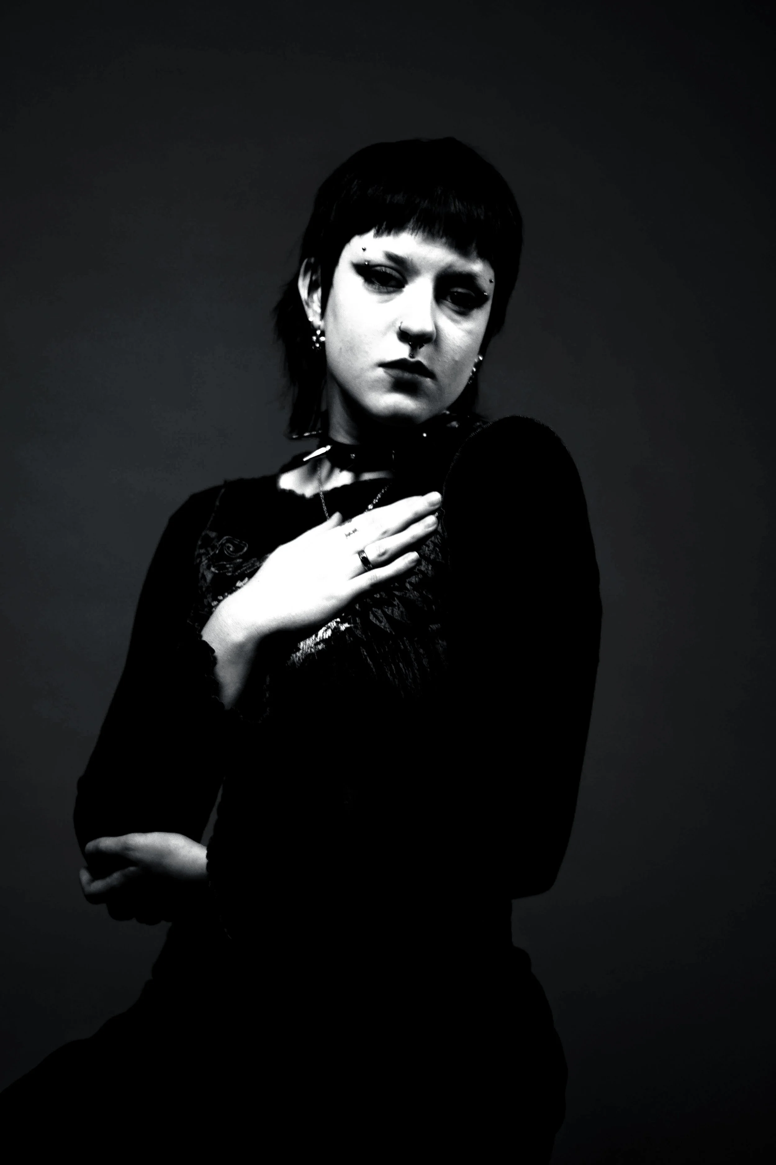 Black and white photo of a woman with short dark hair, multiple facial piercings, and wearing dark clothing and jewelry, looking directly at the camera with one hand on her chest and the other arm crossed.