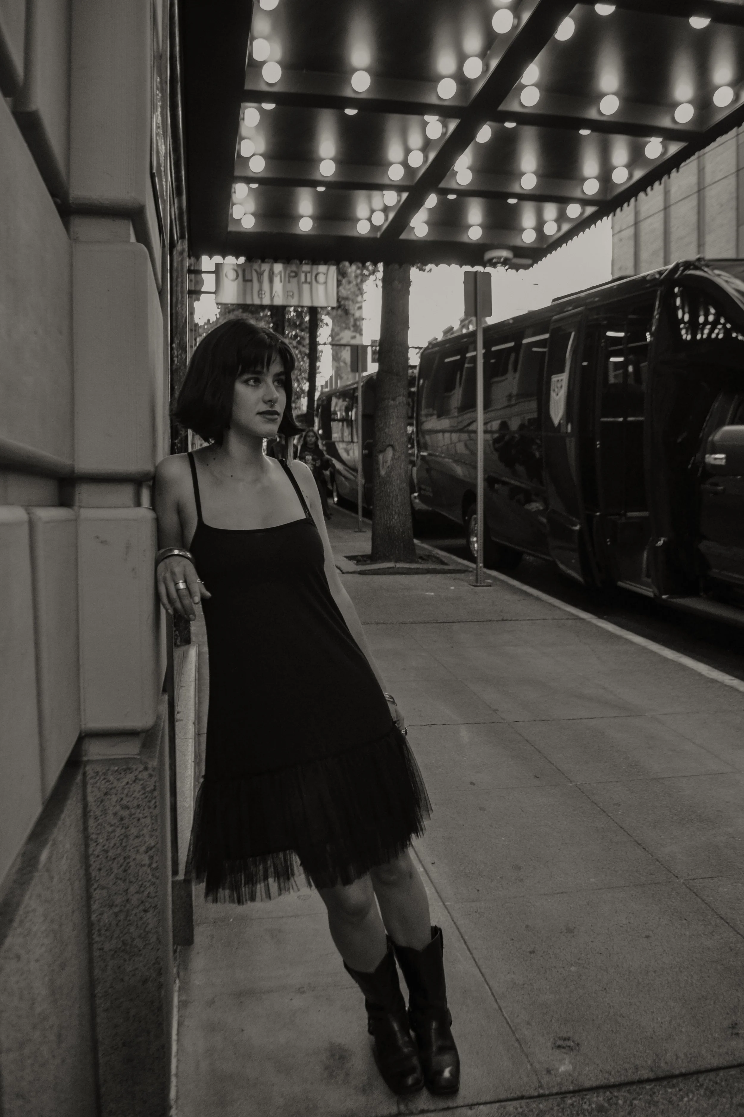 A young woman with dark hair and tattoos wearing a black dress and boots, leaning against a wall outside a bar called Olympic Bar, with buses and trees in the background.