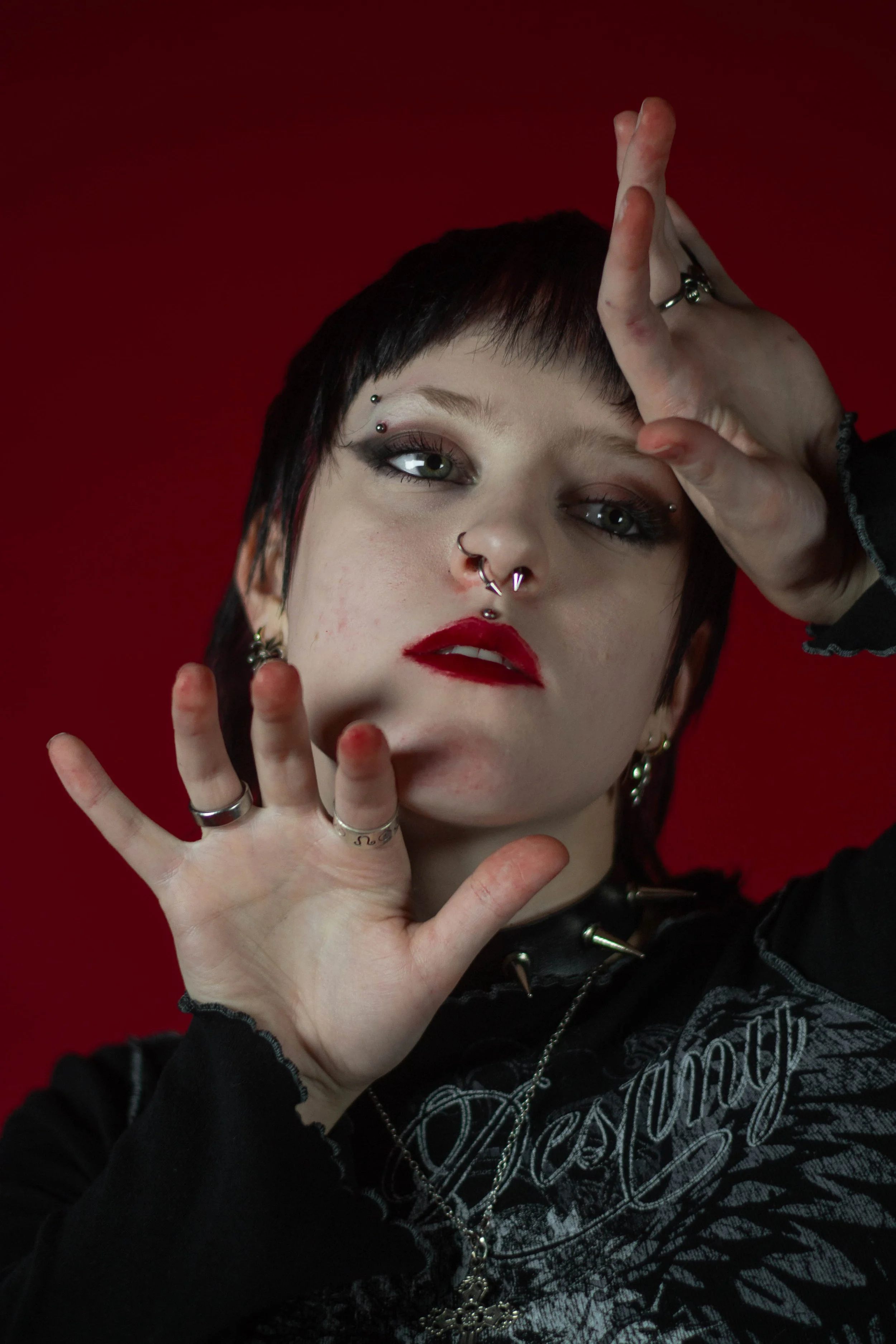 A person with short dark hair, multiple facial piercings, makeup, and various rings holding their hand up near their face against a red background.