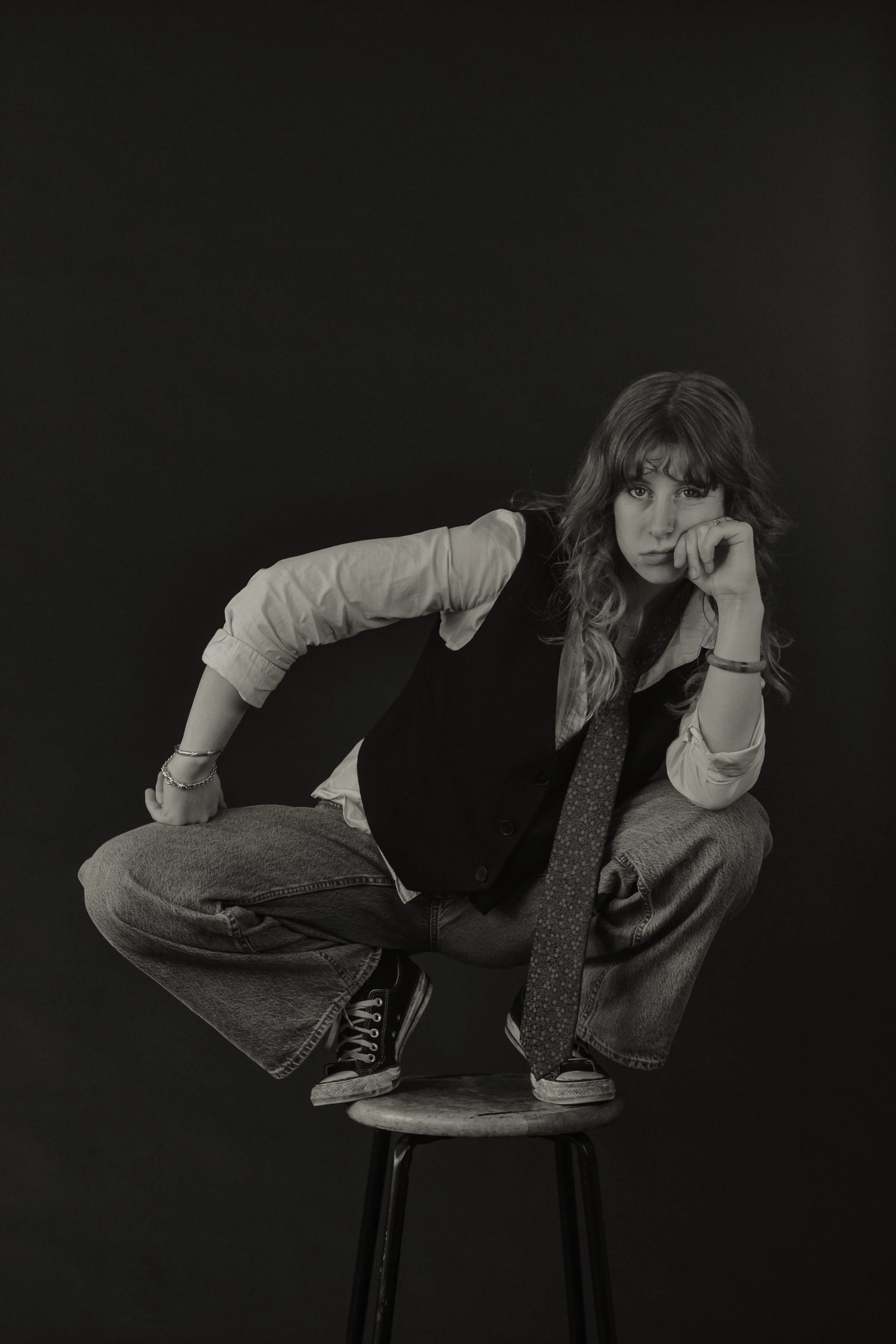 A young woman with curly hair sitting on a stool with black background, in a pose with one knee on the stool, leaning forward, resting her chin on her hand, wearing casual clothing including a vest, shirt, and sneakers.