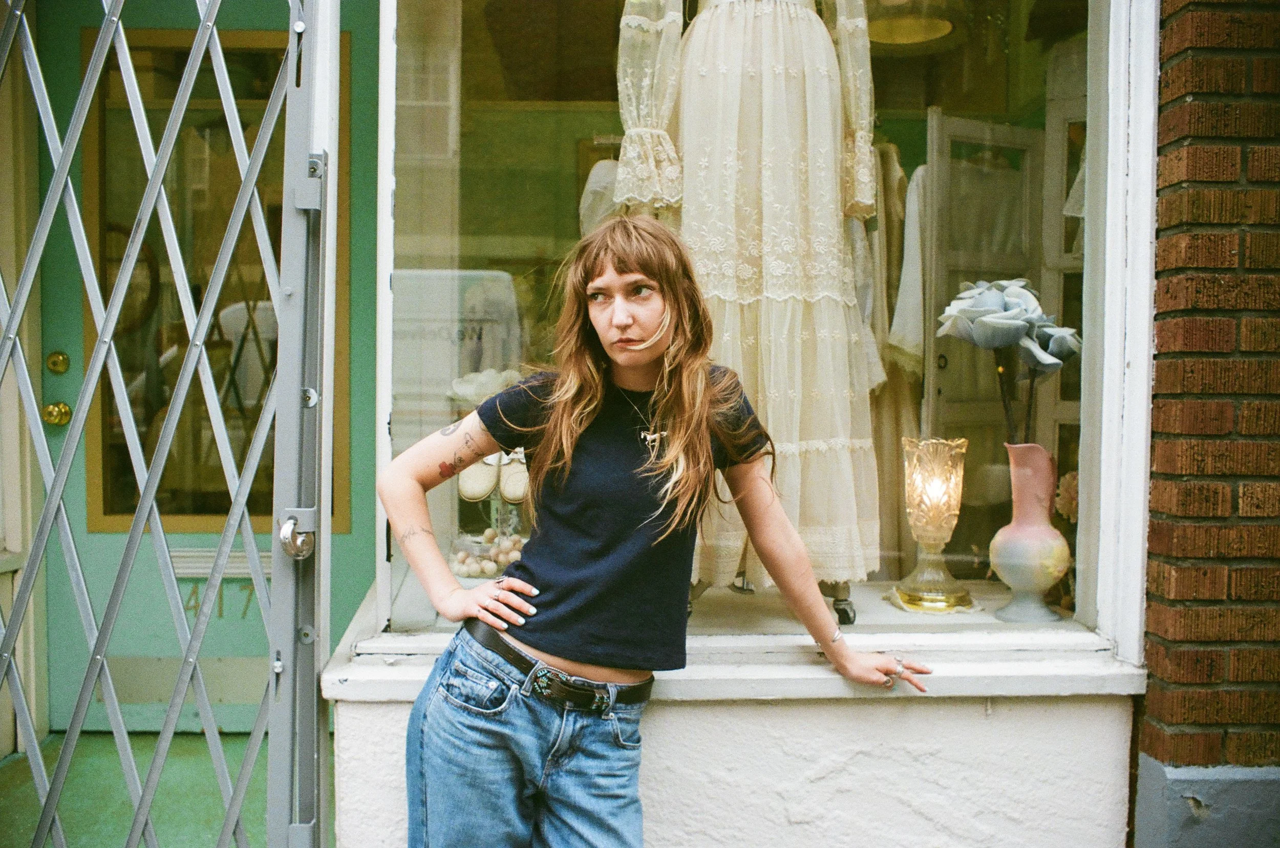 A young woman with long wavy hair, wearing a black T-shirt and blue jeans, standing in front of a storefront window display with vintage clothing and accessories. She has a tattoo on her left arm and is posing with one hand on her hip, looking slight