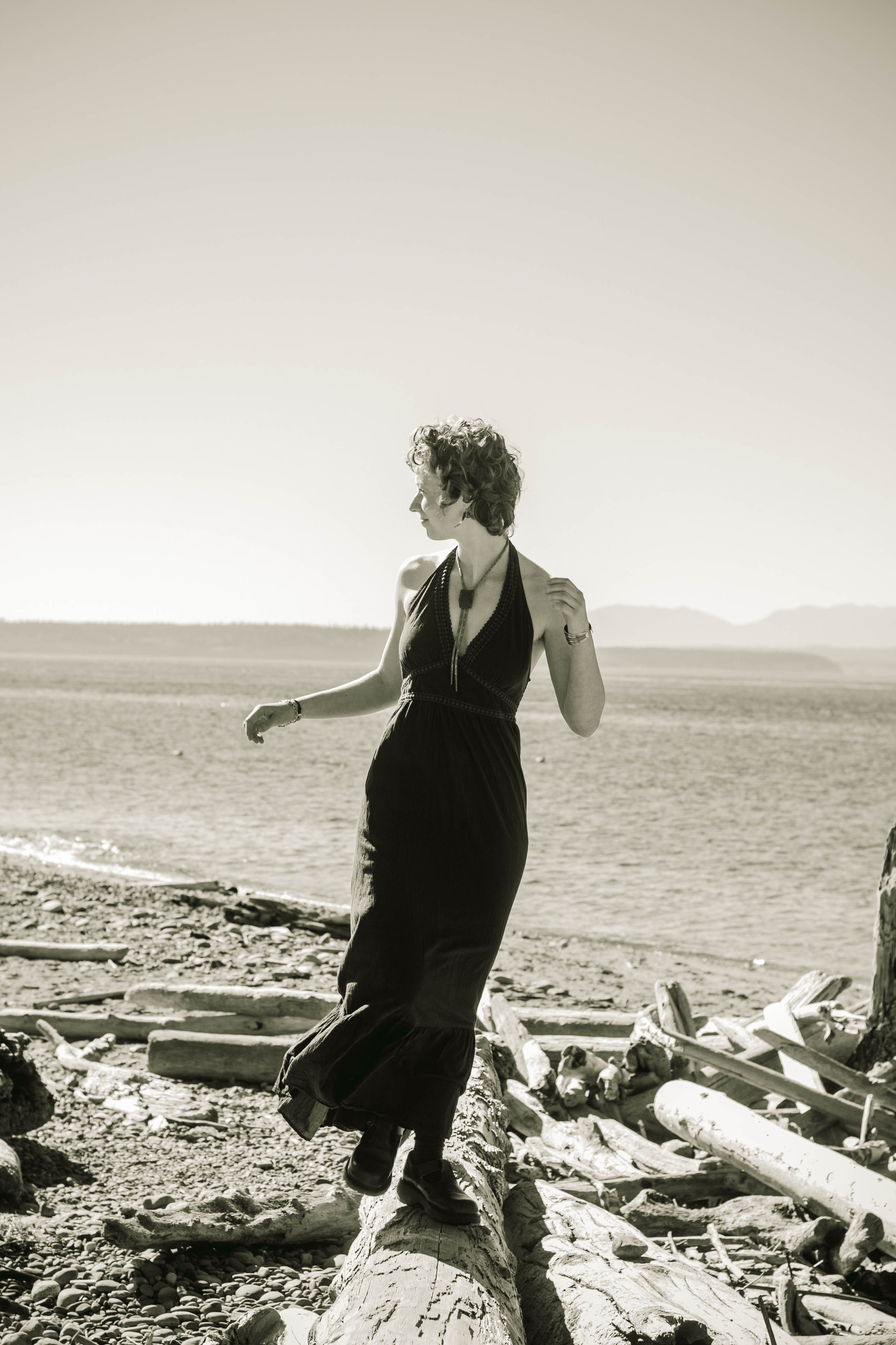 A woman with curly hair wearing a long dress and jewelry, standing on a log on a beach with driftwood, looking to the side.