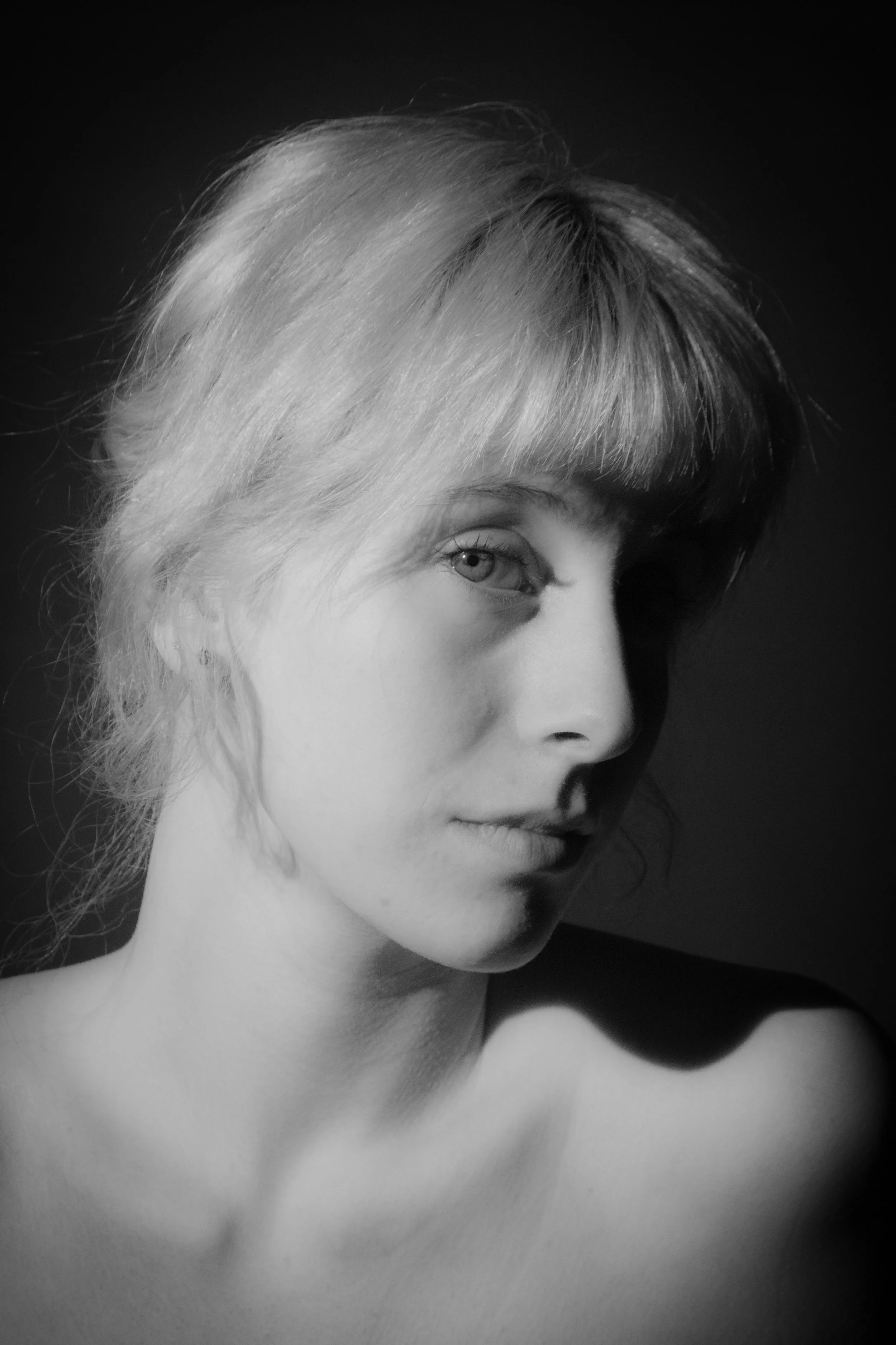 Black and white portrait of a woman with light-colored hair and a side profile, showing her face, neck, and shoulder against a dark background.